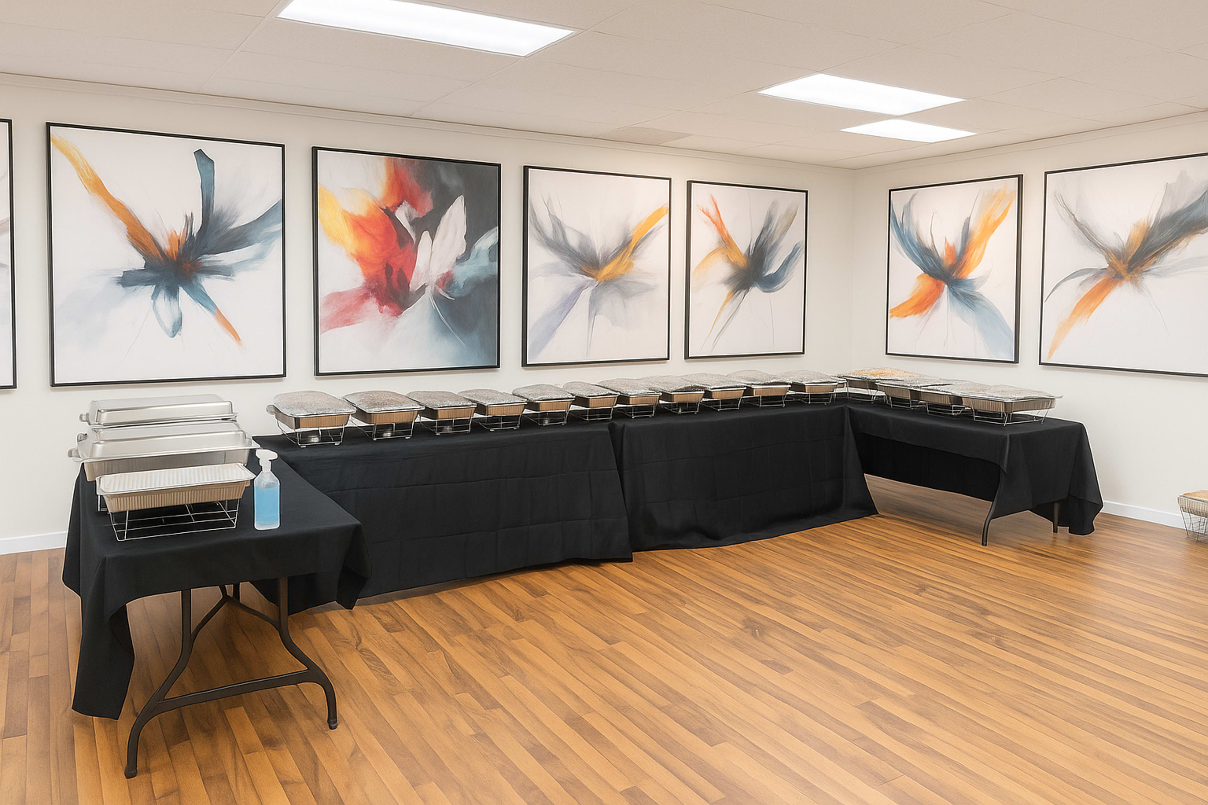 A buffet table is set up against a wall adorned with abstract artwork, featuring covered food trays and a hand sanitizer station nearby.