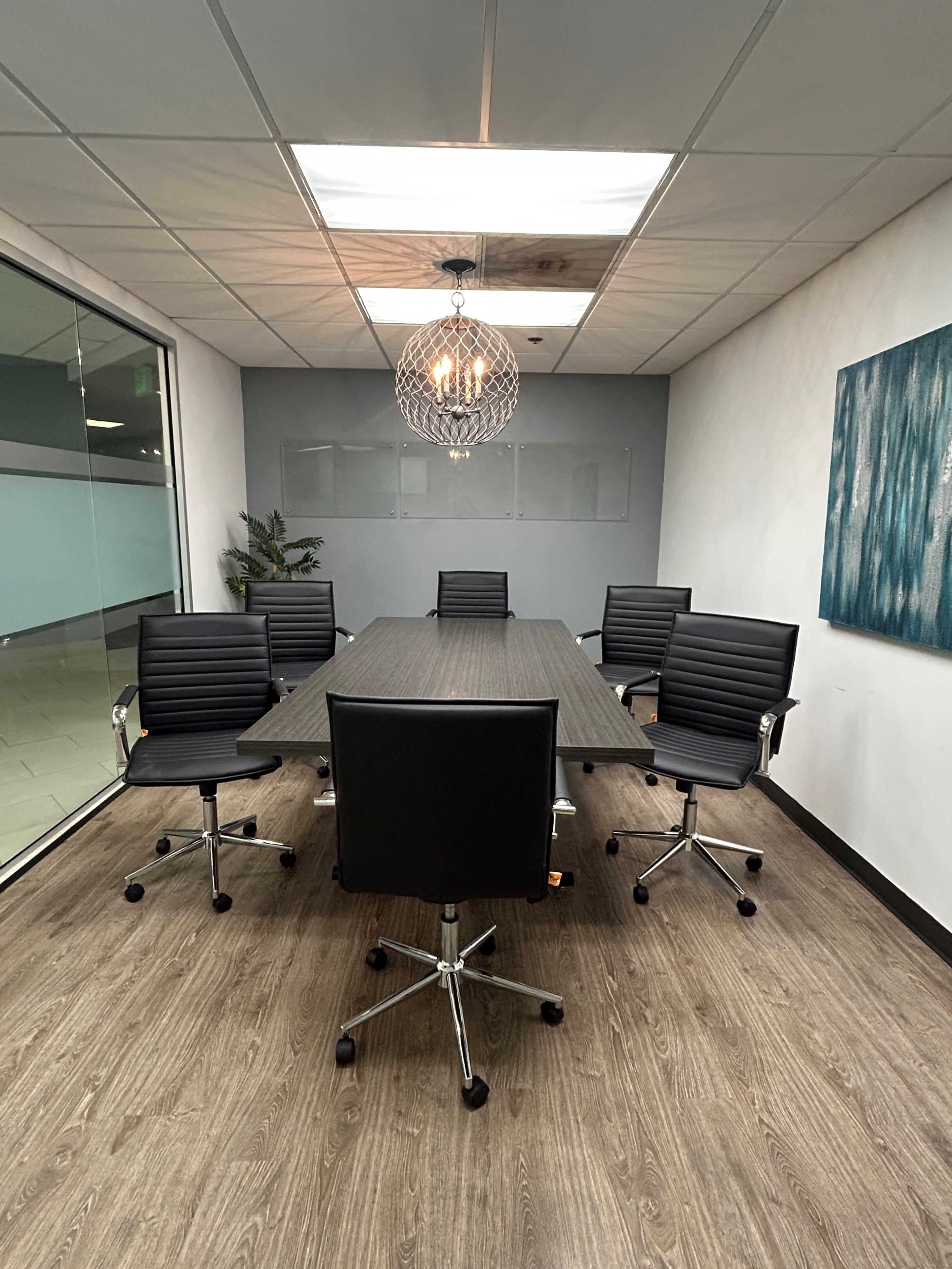 A conference room features a long table surrounded by six black rolling chairs under a modern light fixture.