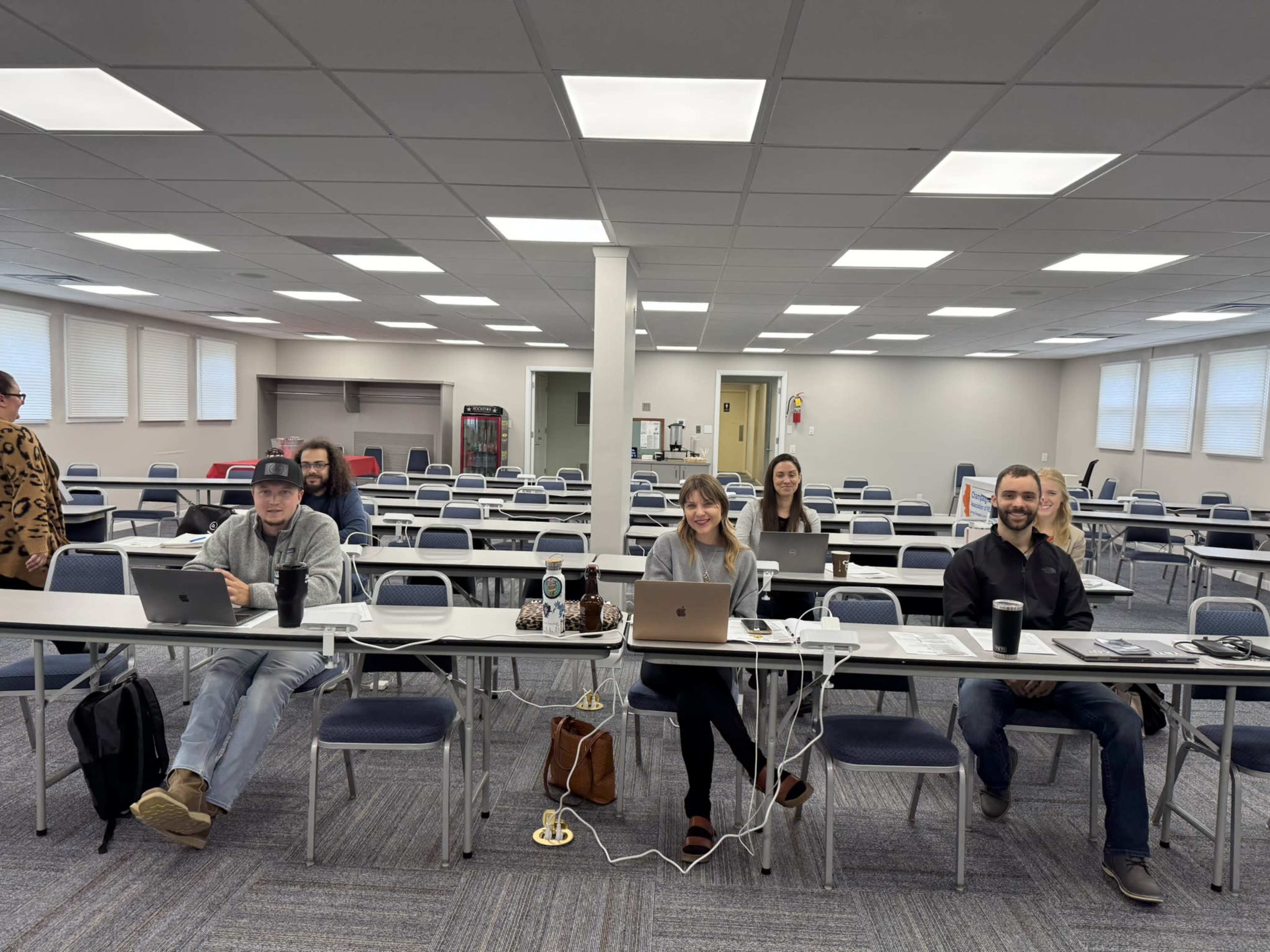 A group of five people is seated at long tables in a classroom setting, each working on laptops, while several empty chairs are visible in the background.