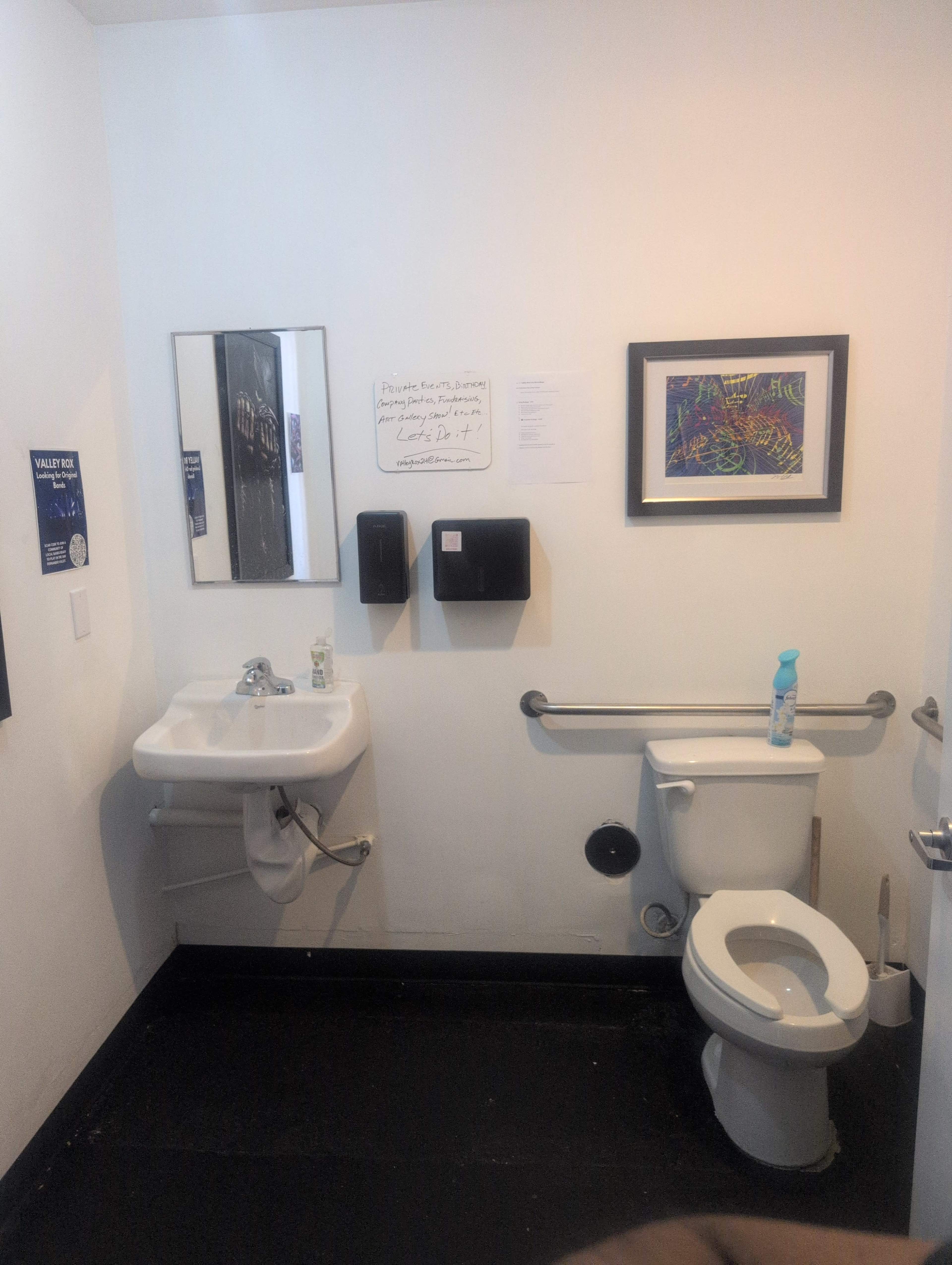 The image shows a small restroom with a sink, mirror, toilet, and wall-mounted soap dispensers, along with an art piece on the wall.