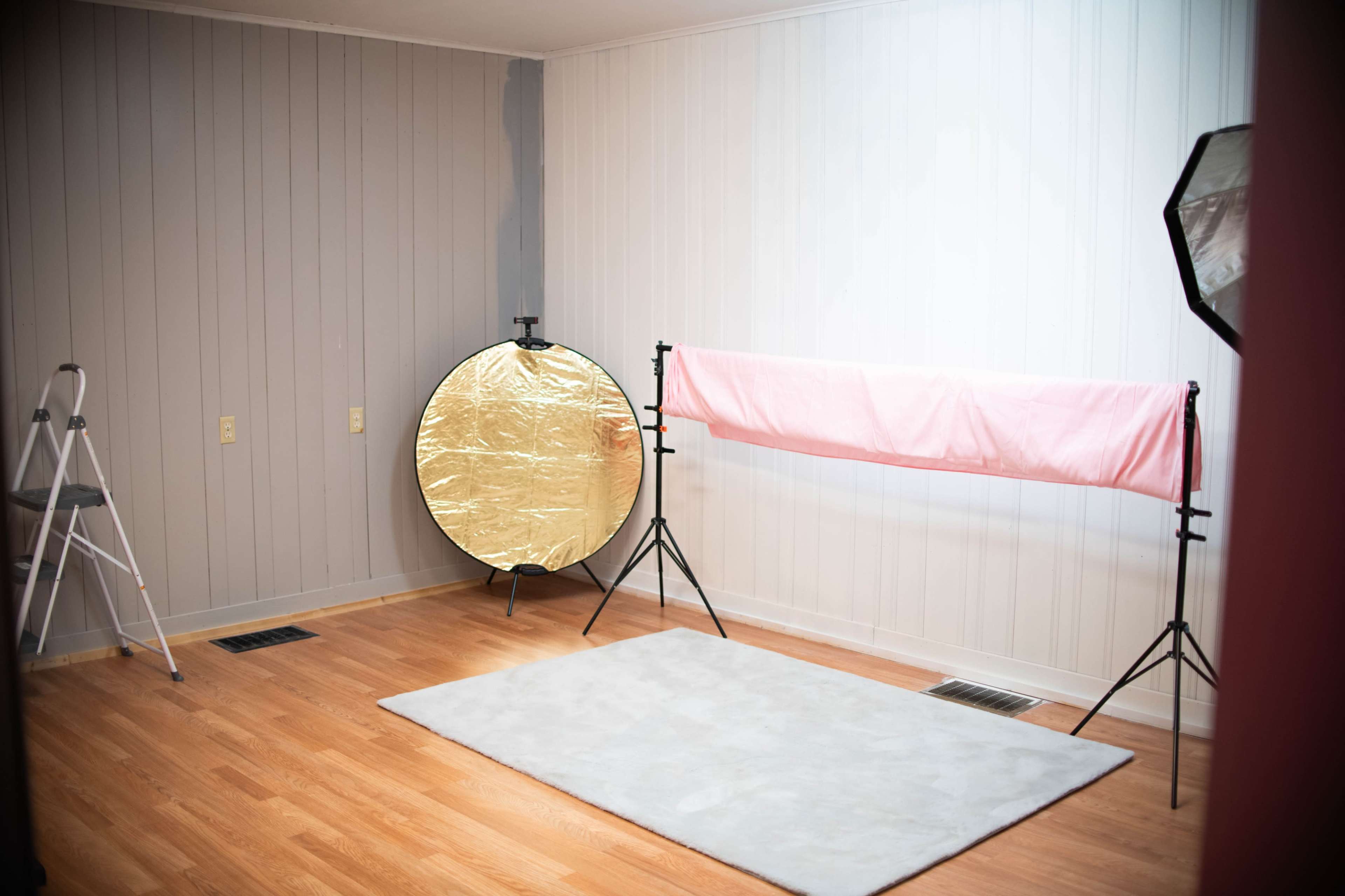 The image shows a photography studio with a gray wall, wooden flooring, a pink backdrop draped over a stand, a large round reflector, and a step ladder.