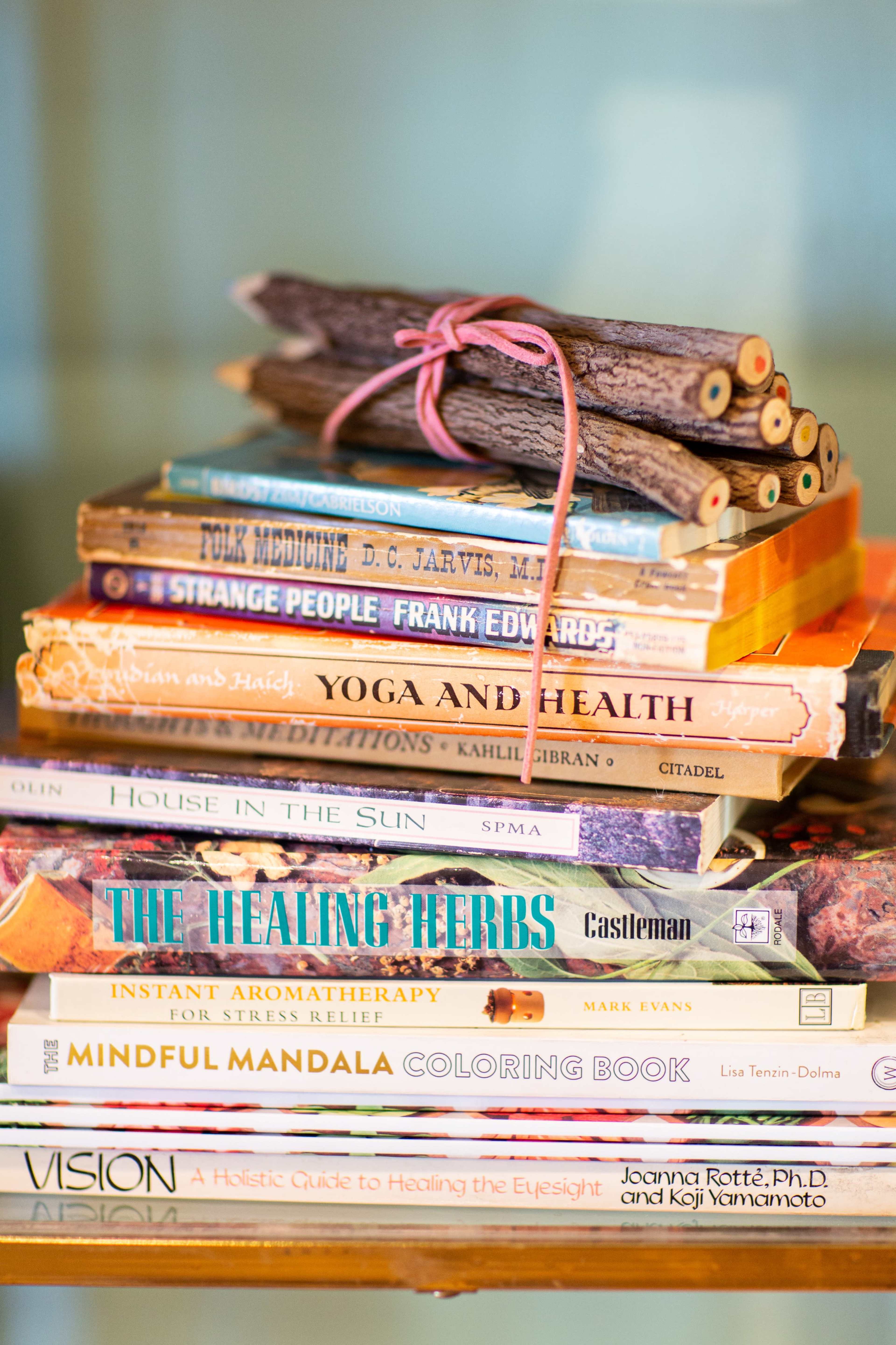 A stack of various books on health, healing, and mindfulness is topped with a bundle of small sticks tied with string.
