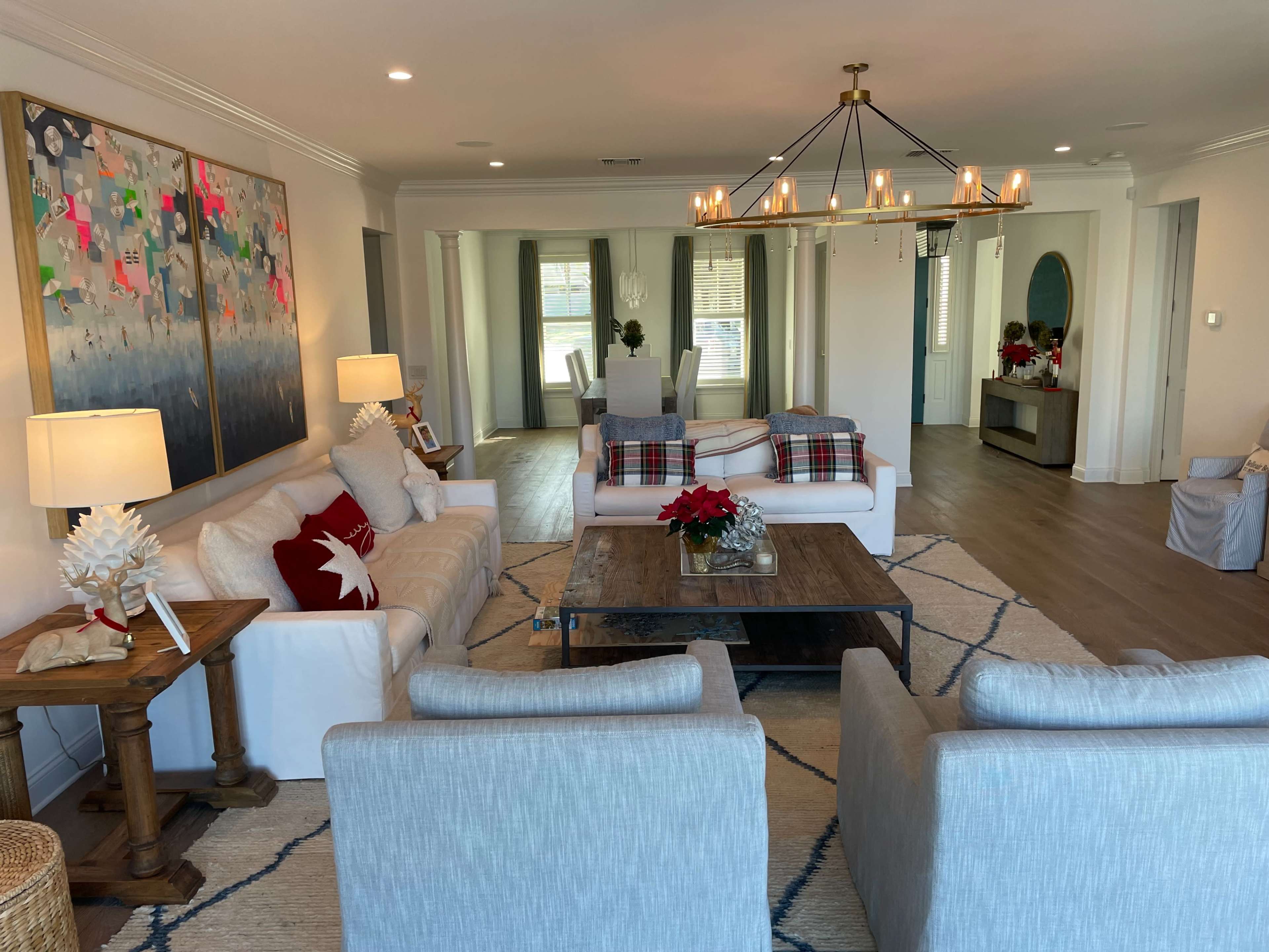 A living room furnished with a coffee table, two light gray armchairs, a white couch with decorative pillows, and artwork on the walls, illuminated by a large chandelier.