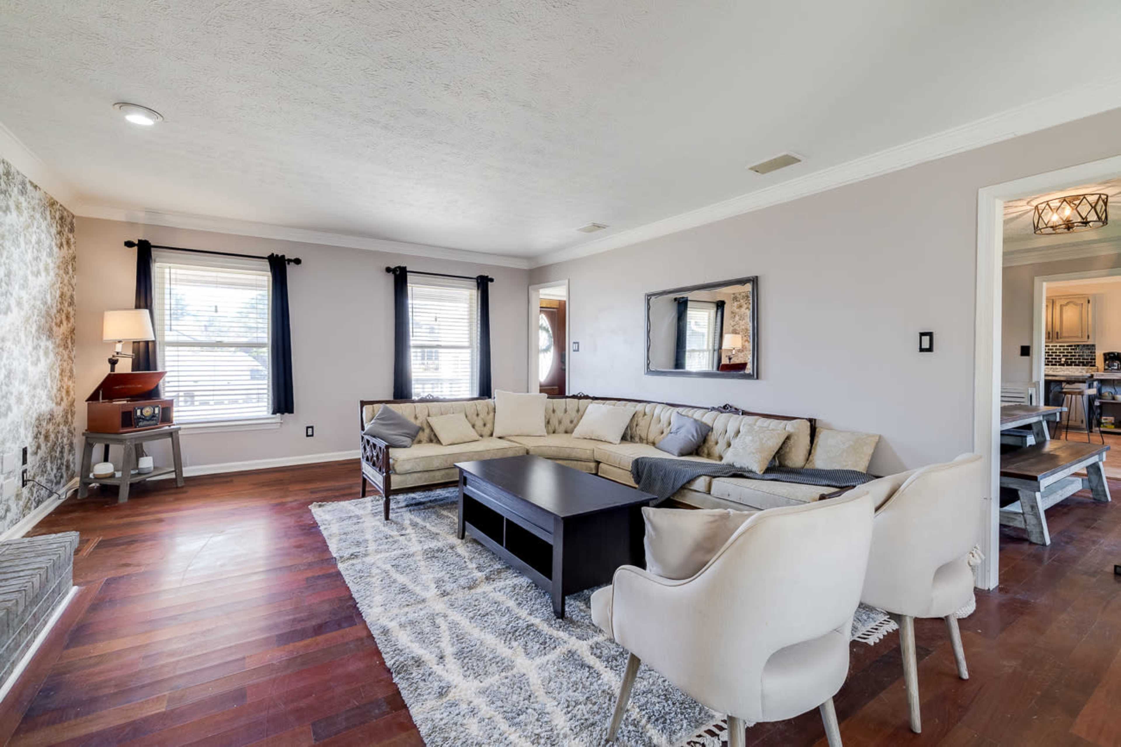 A spacious living room featuring a large sectional sofa, a coffee table, and windows with black curtains, complemented by hardwood flooring and a decorative area rug.
