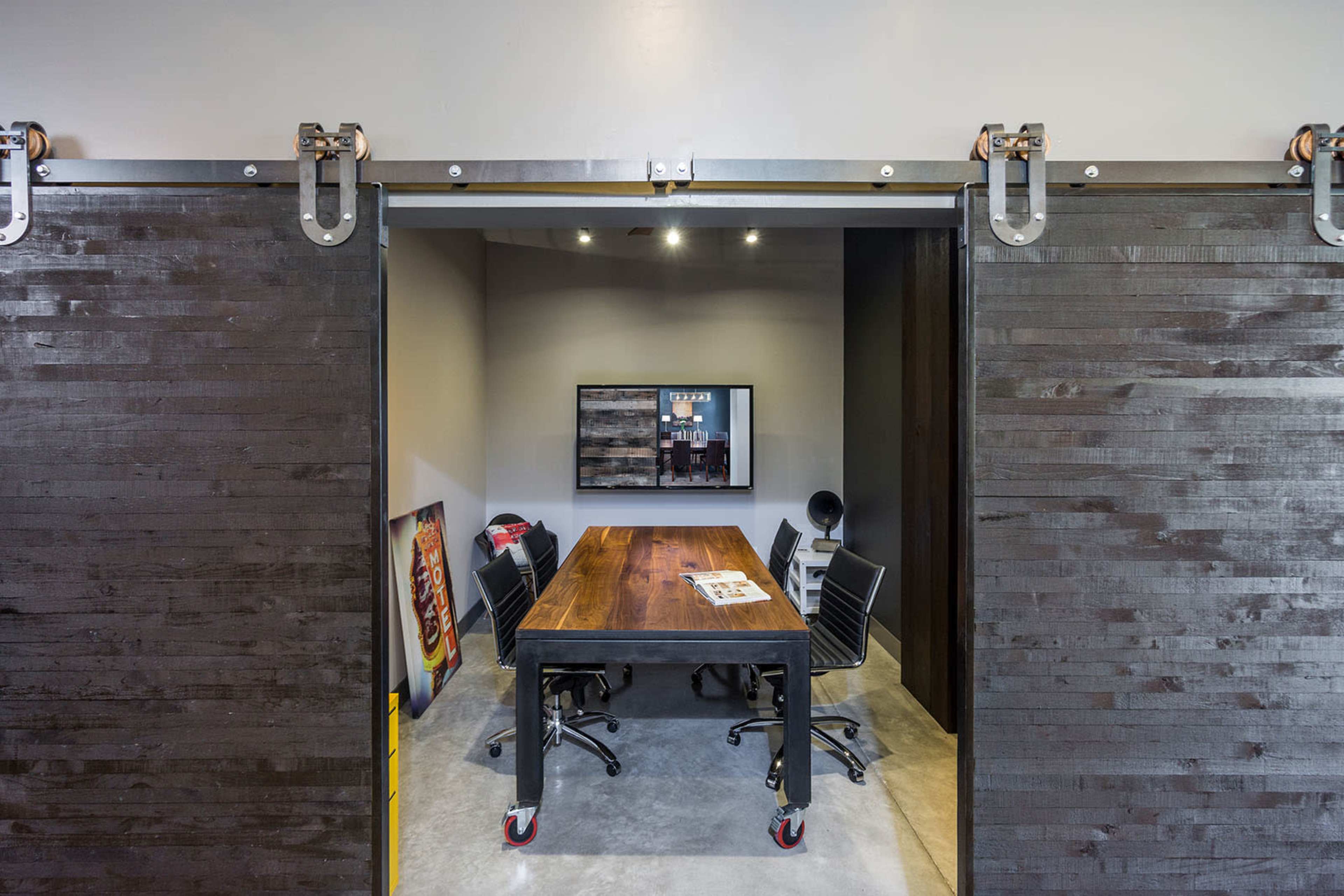 A modern office with a large wooden conference table surrounded by black mesh chairs, viewed through sliding barn doors.