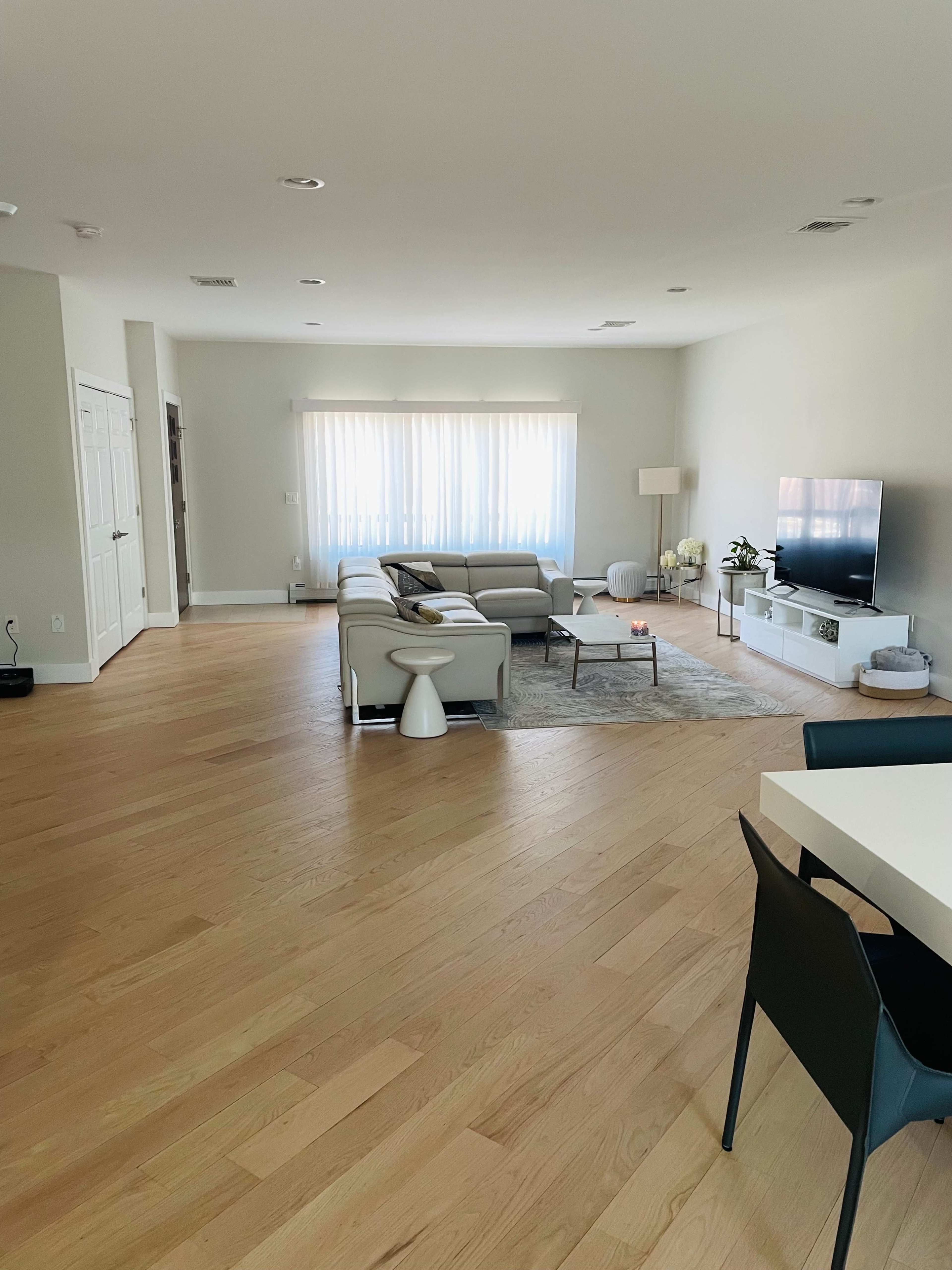 The room features a spacious living area with a light-colored sofa, a television mounted on the wall, and a dining area with chairs on the right.