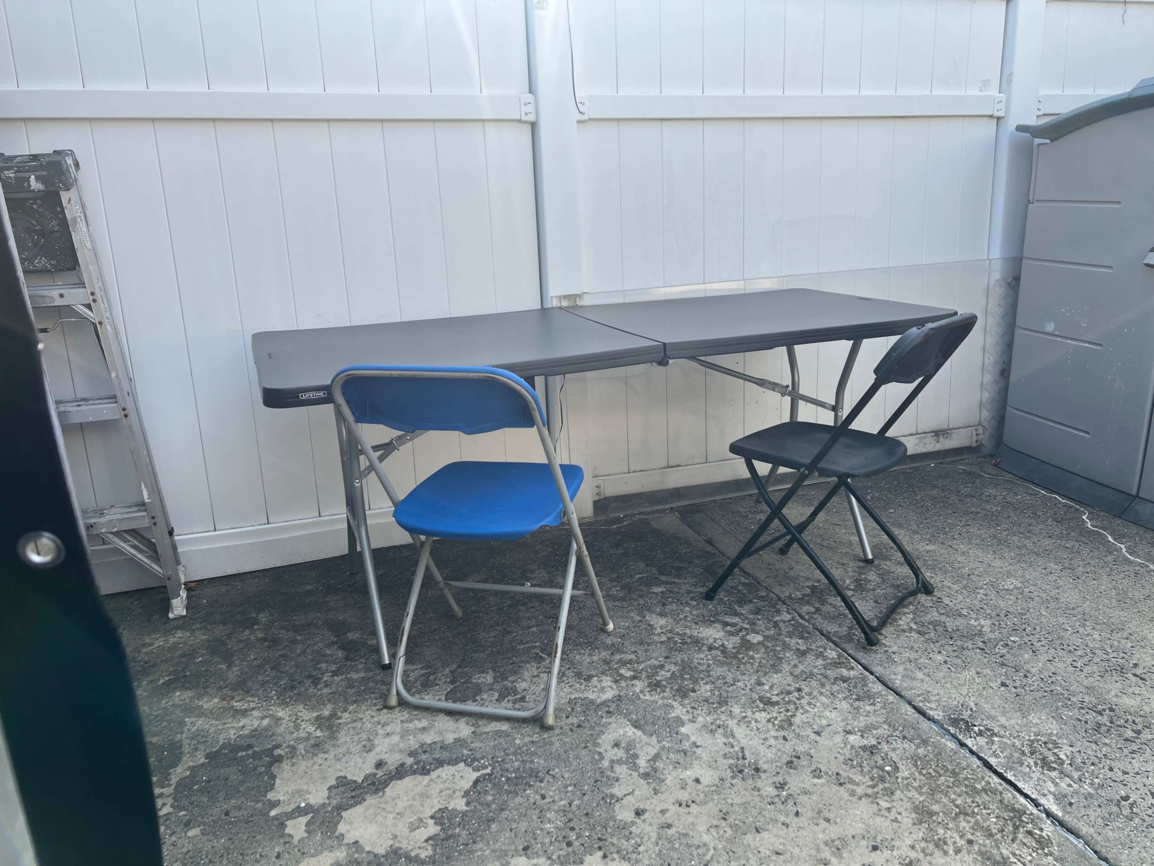 A gray folding table is set up outdoors alongside a blue plastic chair and a black folding chair, with a storage shed visible in the background.
