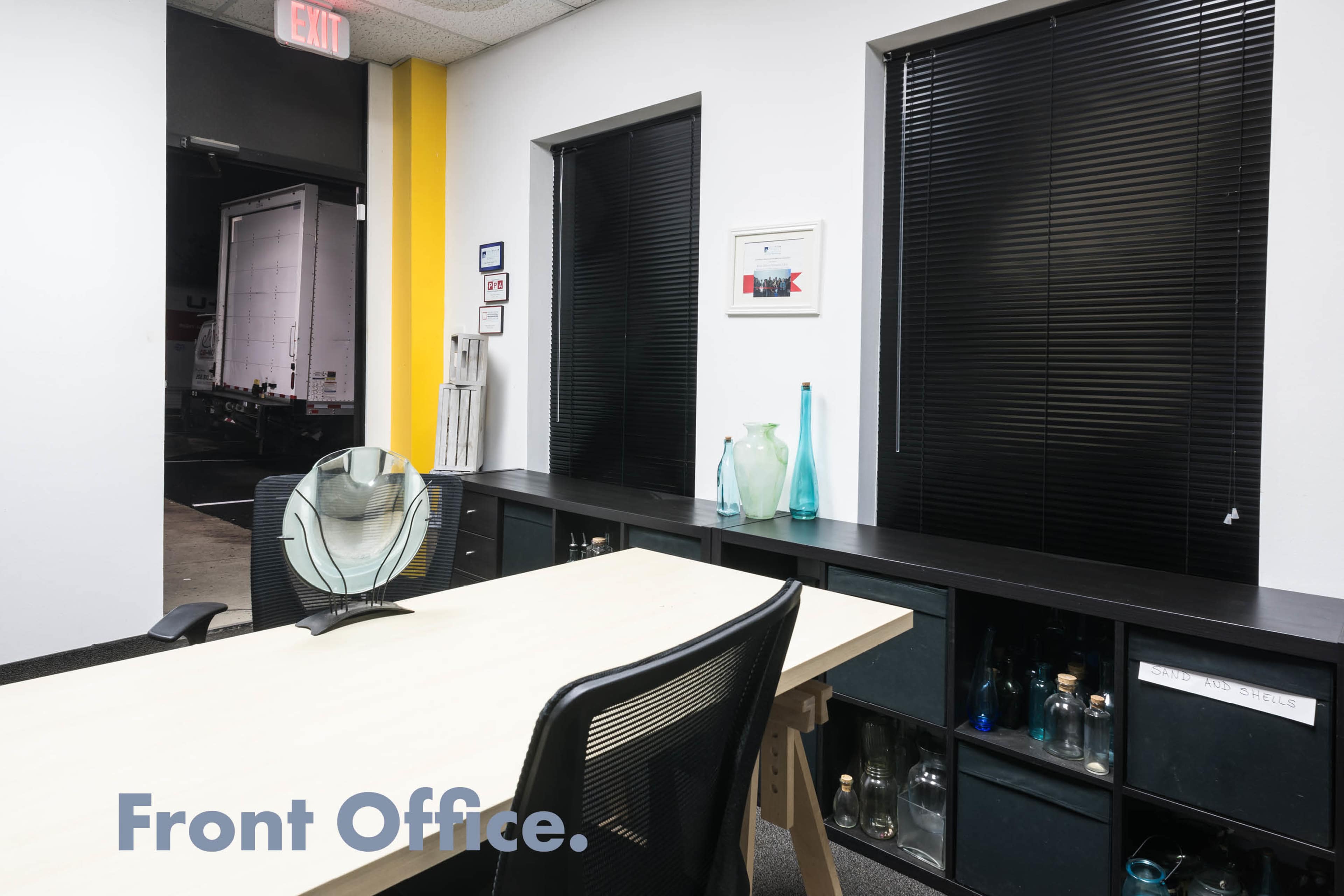 A front office features a desk with a fan and a shelf displaying several decorative glass bottles, alongside a window with blinds partially closed.