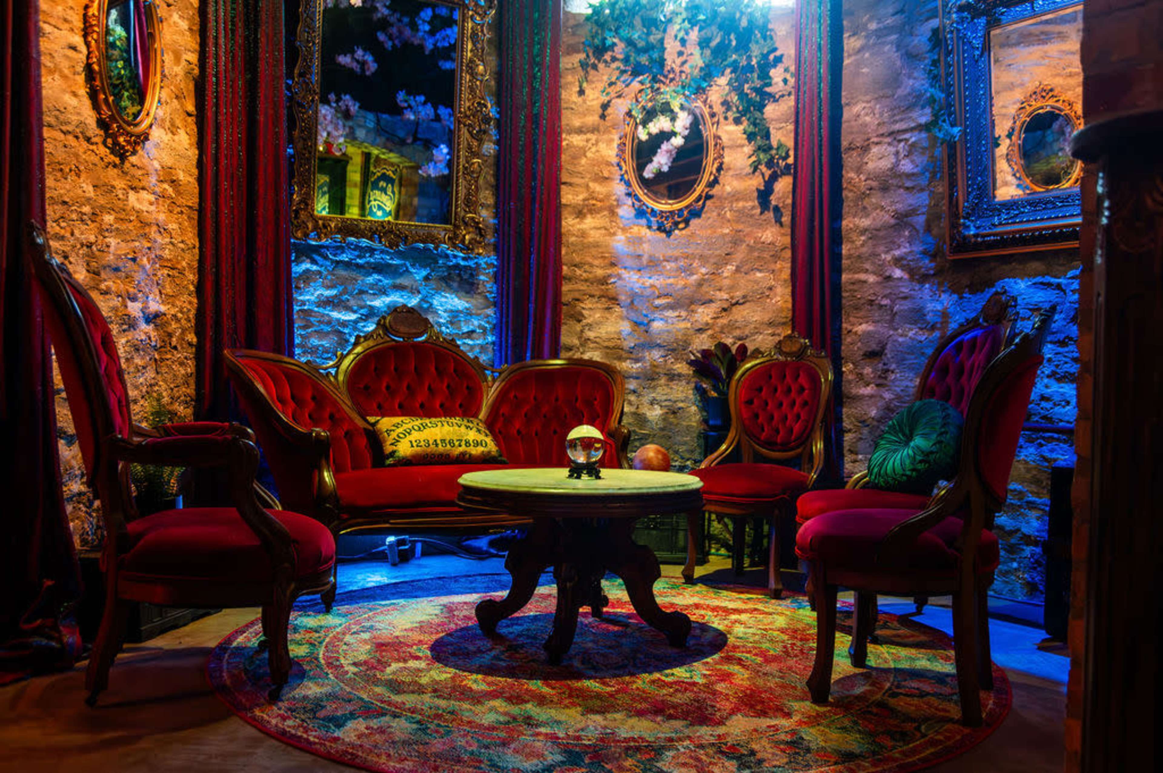The image depicts a cozy parlor with ornate red seating, a round table, and decorative mirrors, all illuminated by colorful lighting.