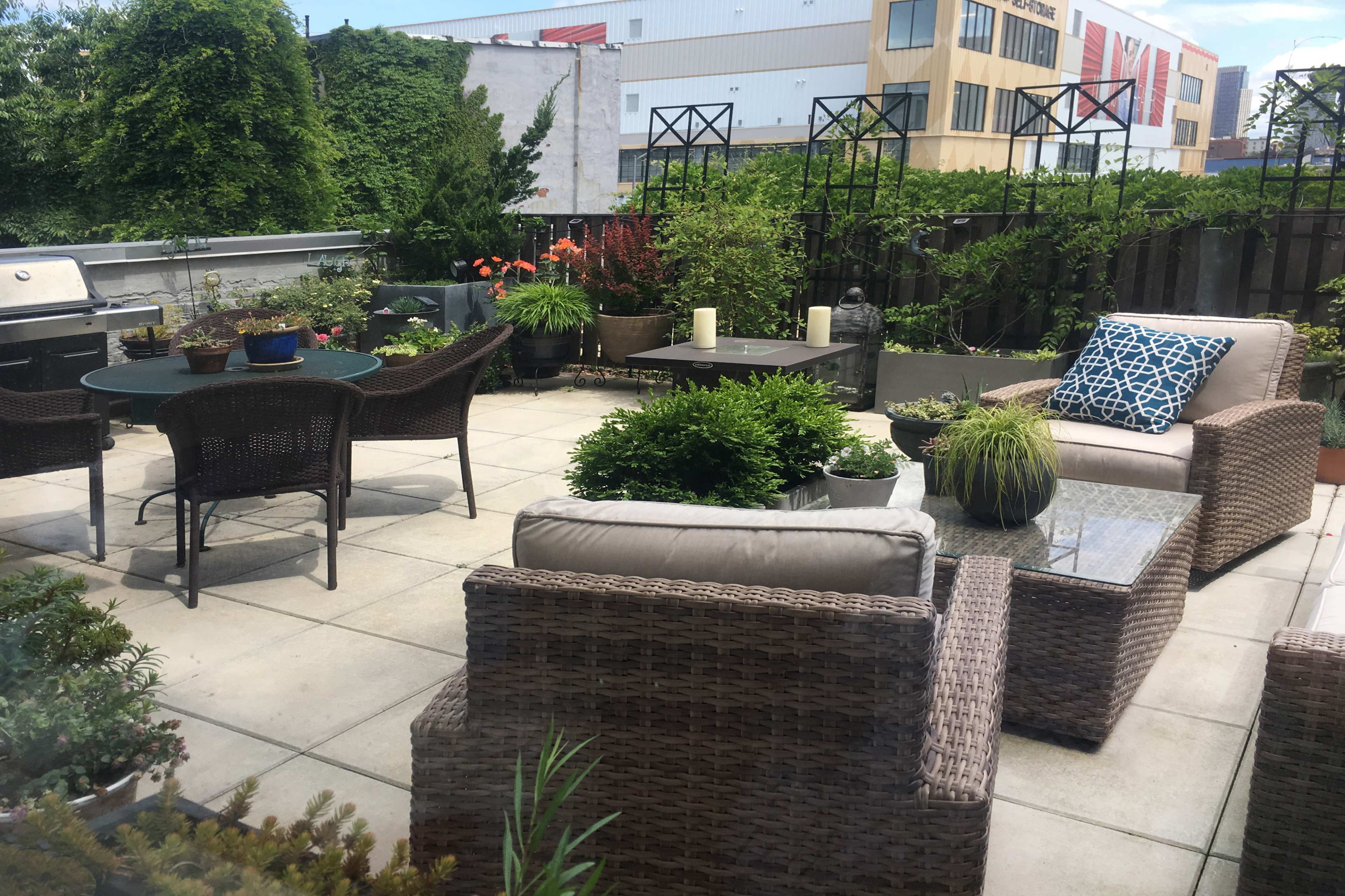 The image shows a patio area with wicker furniture, potted plants, and a dining table surrounded by greenery and urban buildings in the background.
