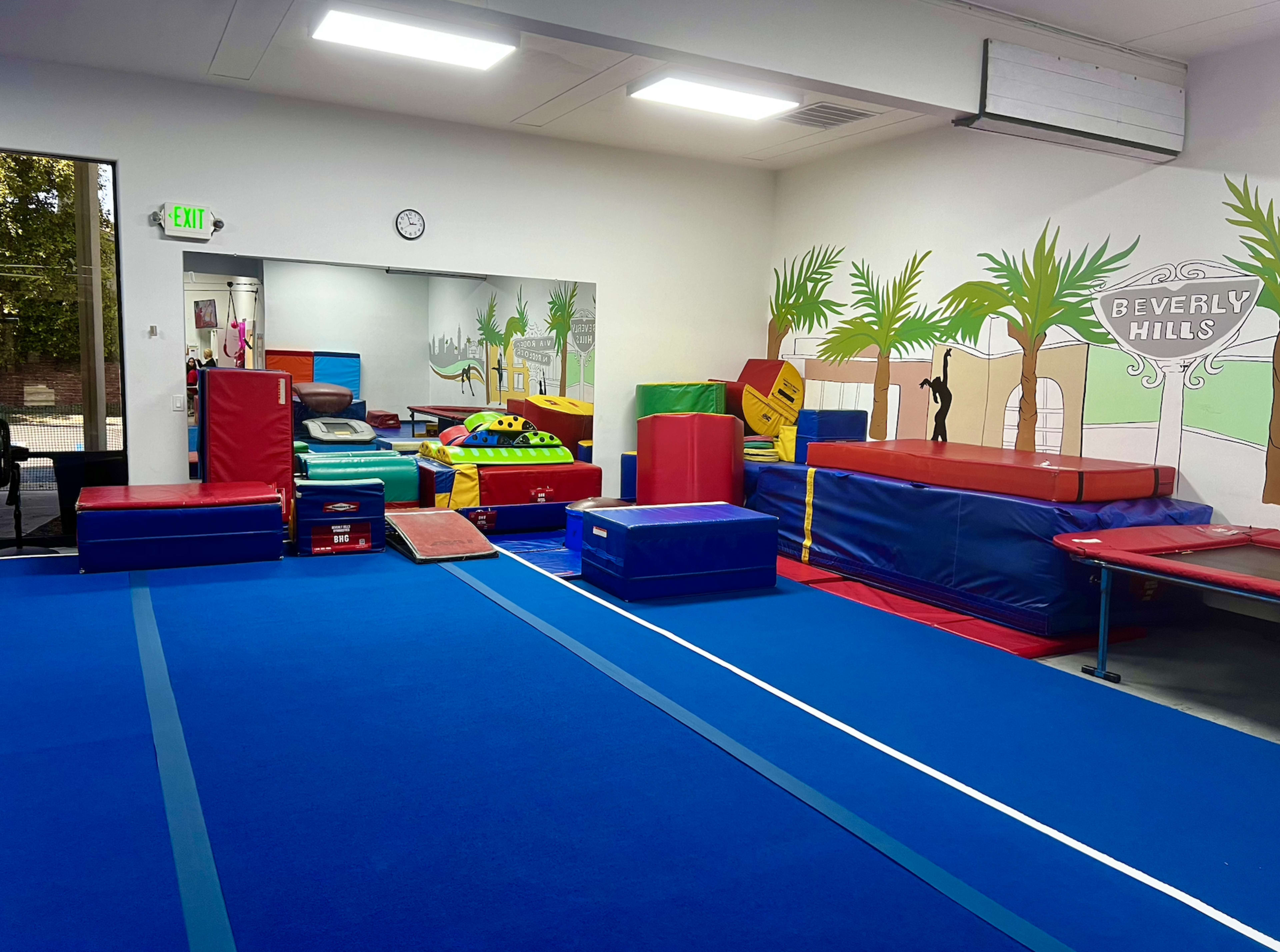 A gymnastic training space with colorful mats, various equipment, and a mirror on one wall, all set against a mural featuring palm trees and "Beverly Hills."
