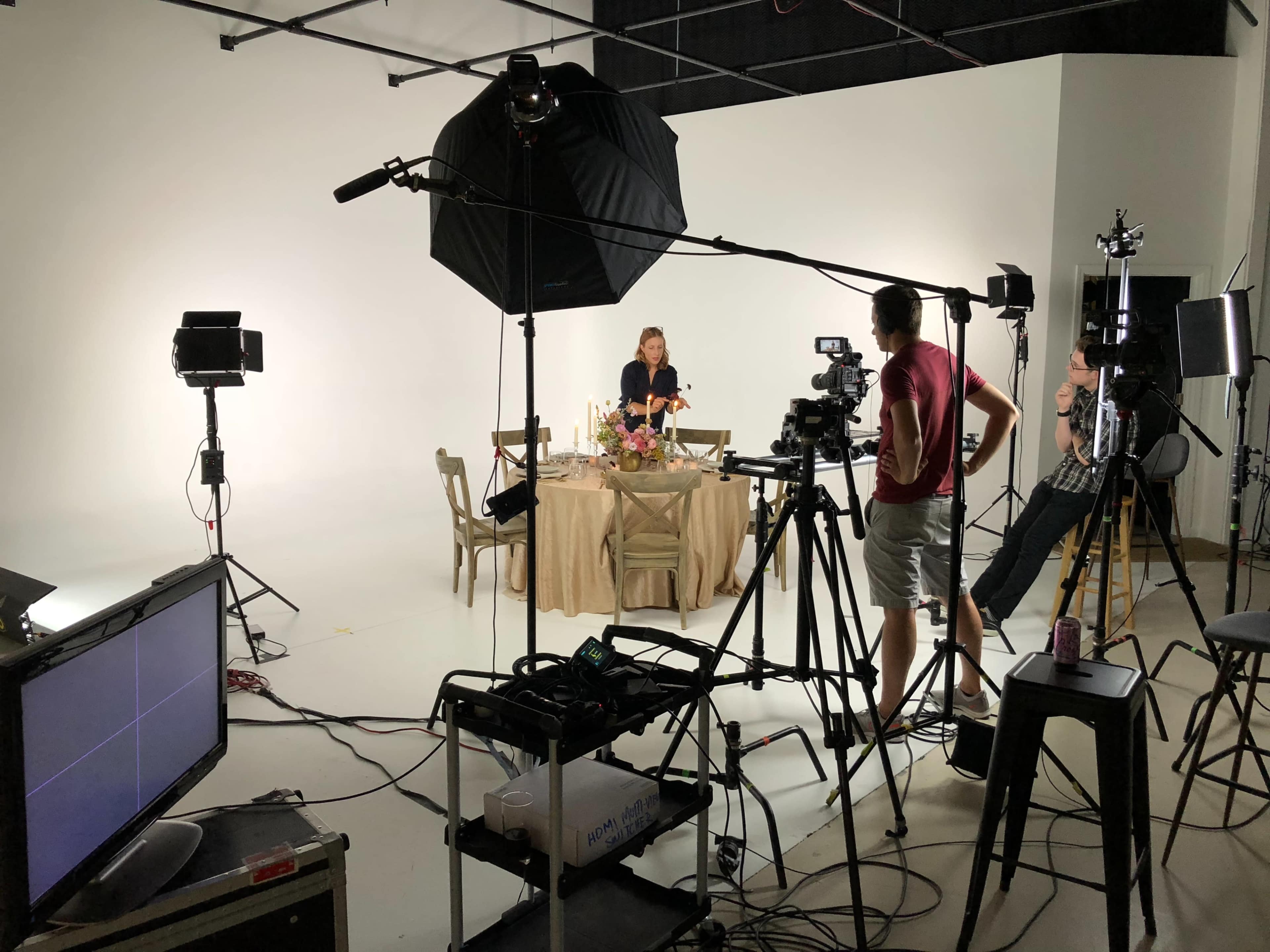 A film crew sets up equipment around a table decorated with flowers in a well-lit studio.