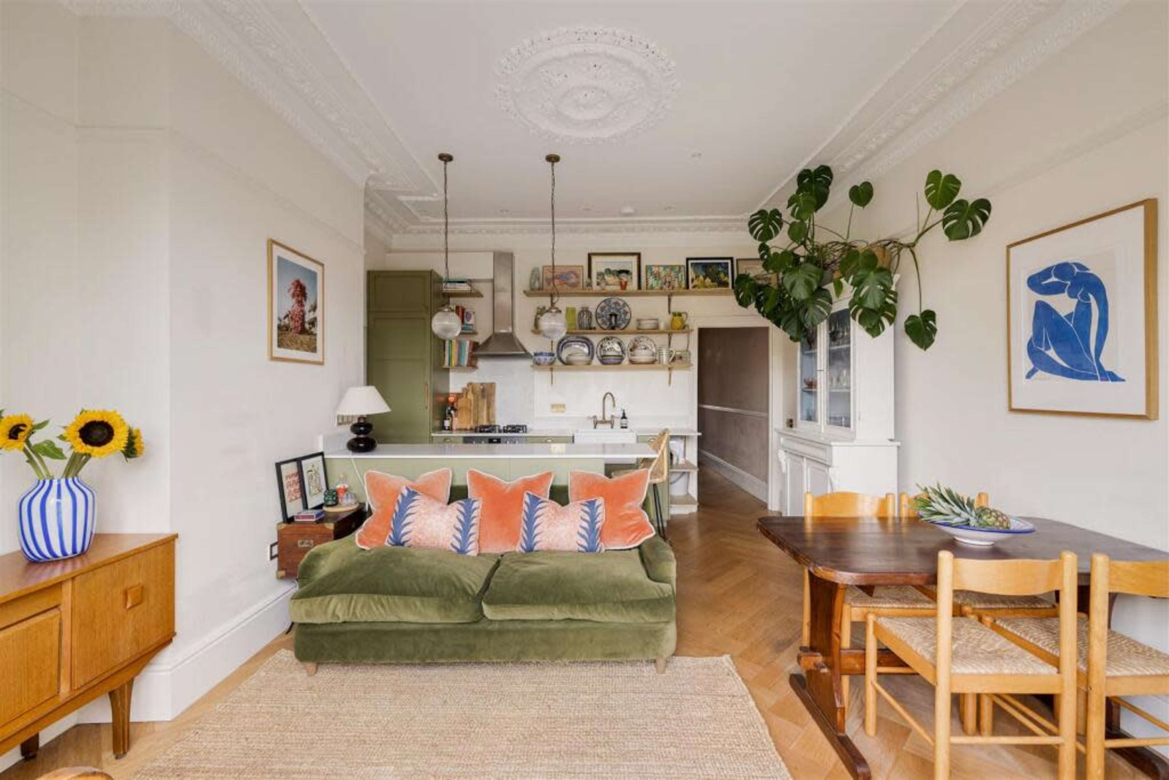 The image shows a bright living space featuring a green sofa with orange and blue cushions, a dining table, and a kitchen area with open shelving and decorative plates.