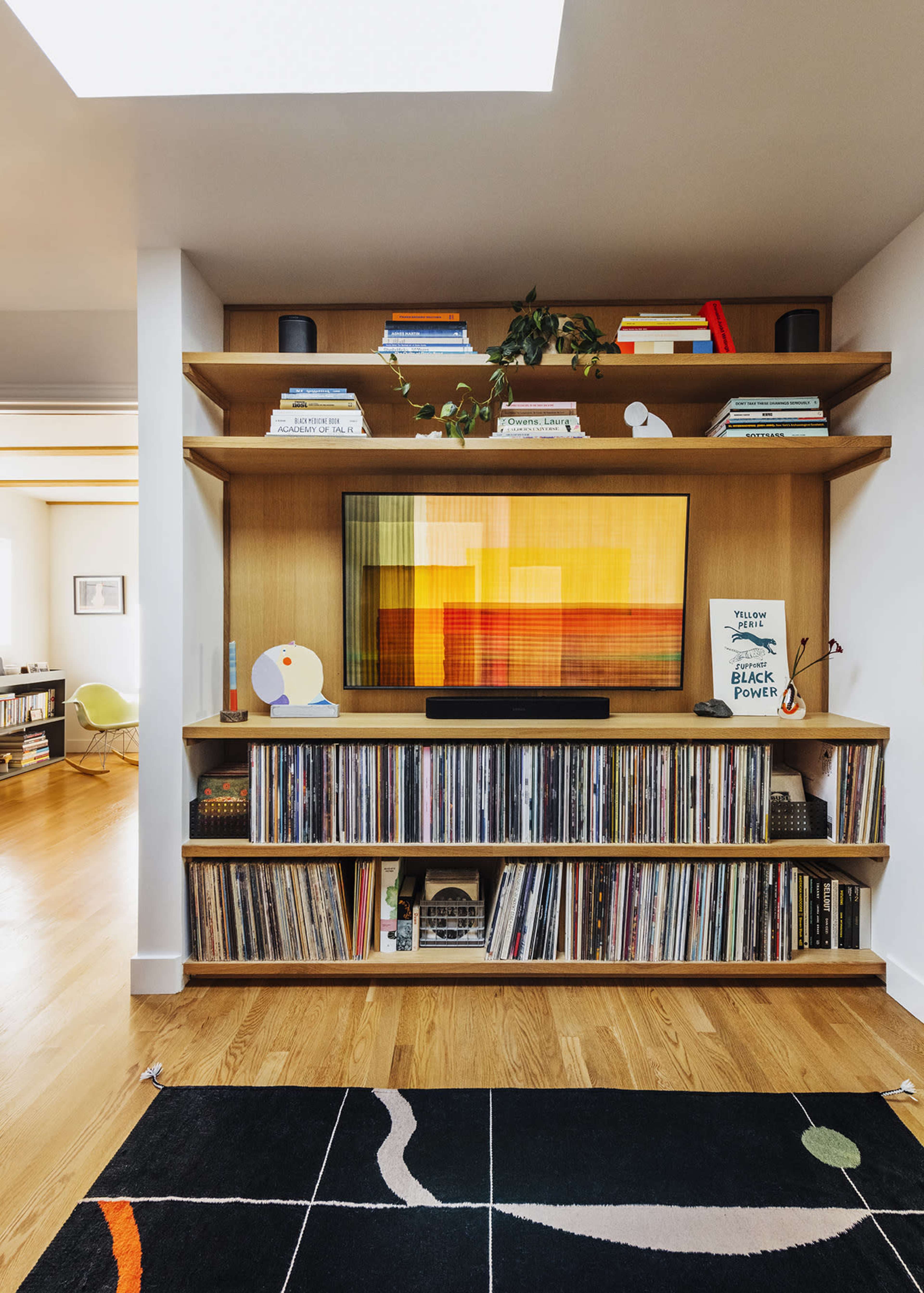The image features a modern living space with a wooden shelf holding books and decorative items above a large television, while a collection of vinyl records is neatly organized below.