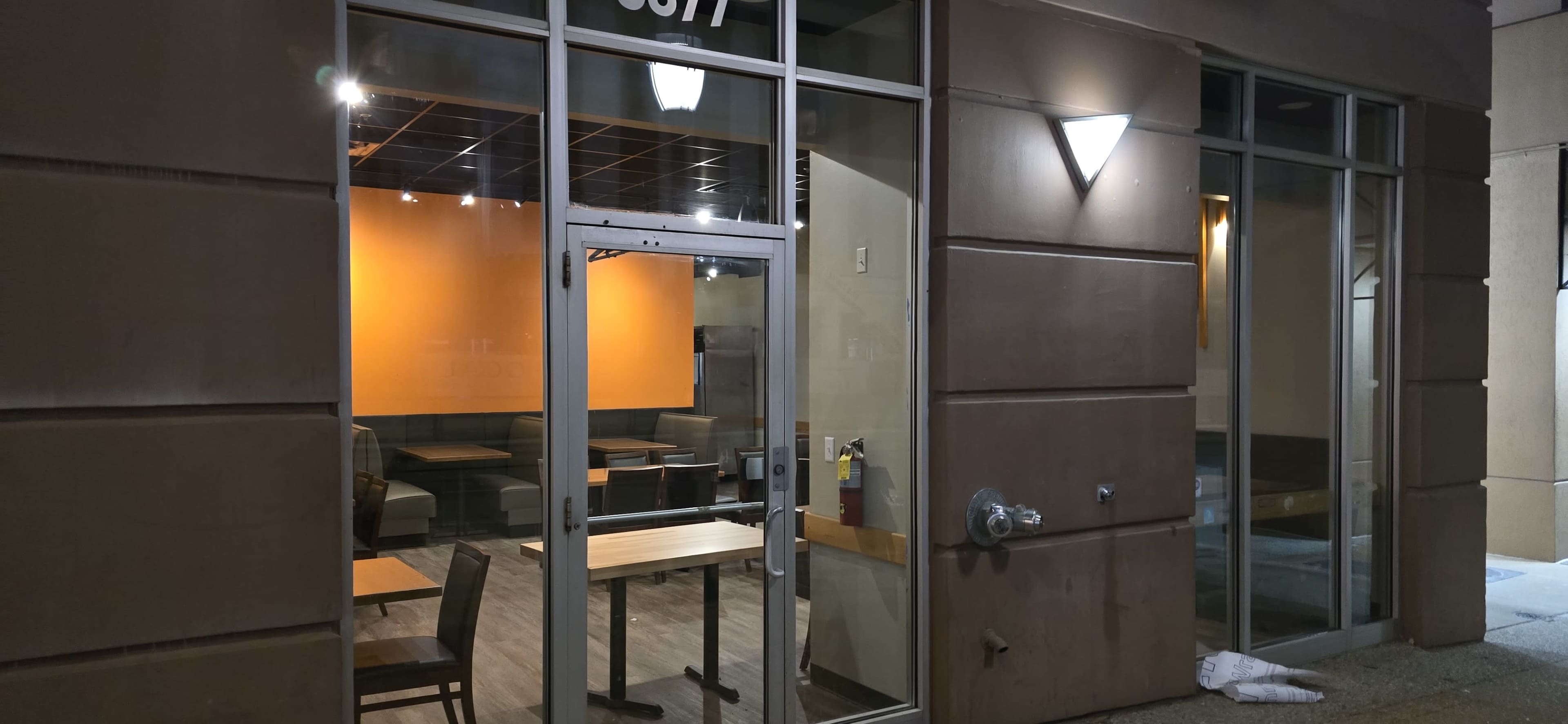 The image shows the exterior of a restaurant with large glass windows and an orange wall visible inside, revealing empty tables and chairs.