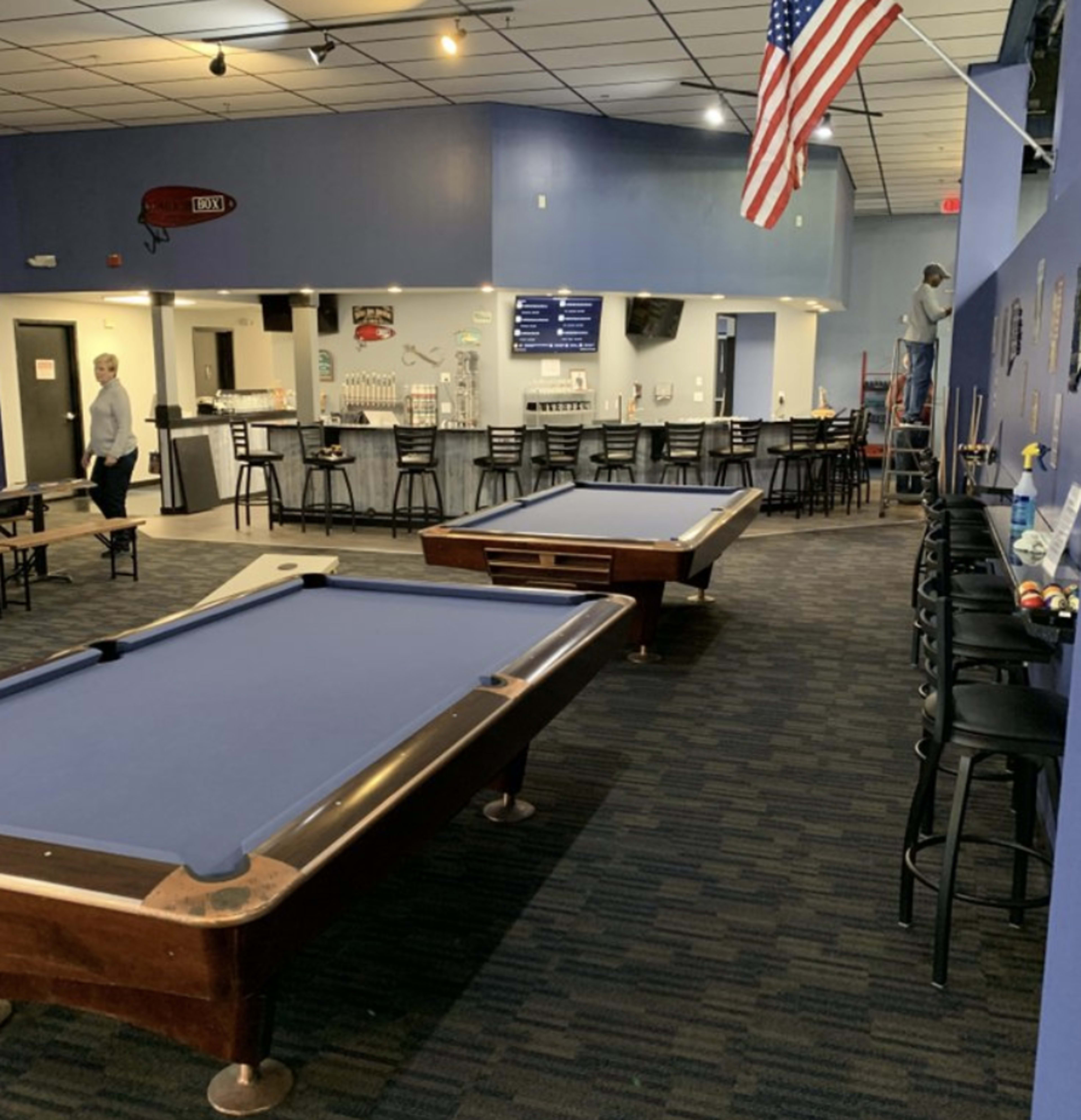 A pool hall with two pool tables and a bar area in the background, featuring an American flag hanging from the ceiling.