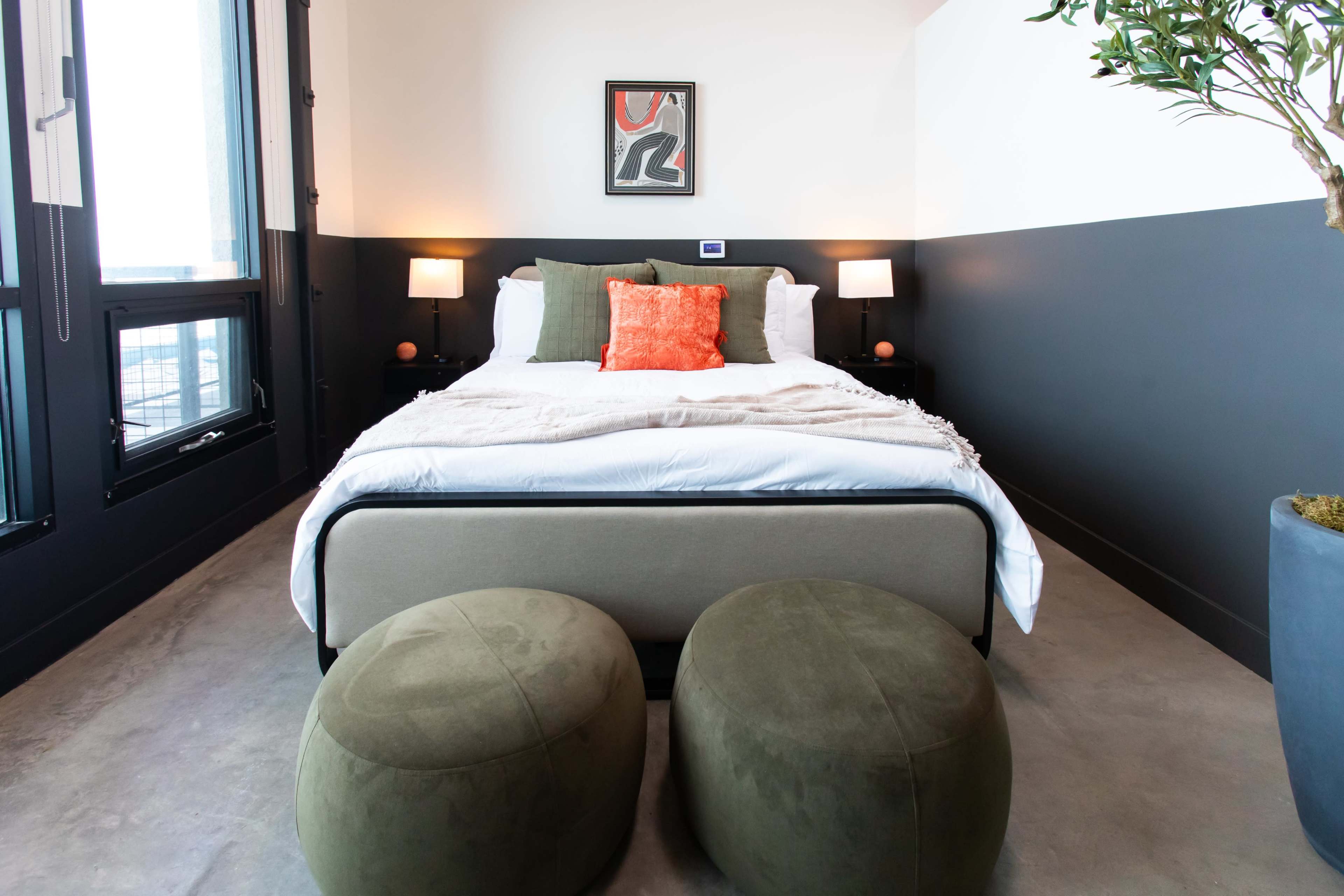 A modern bedroom with a bed, two round green stools, and large windows providing natural light.