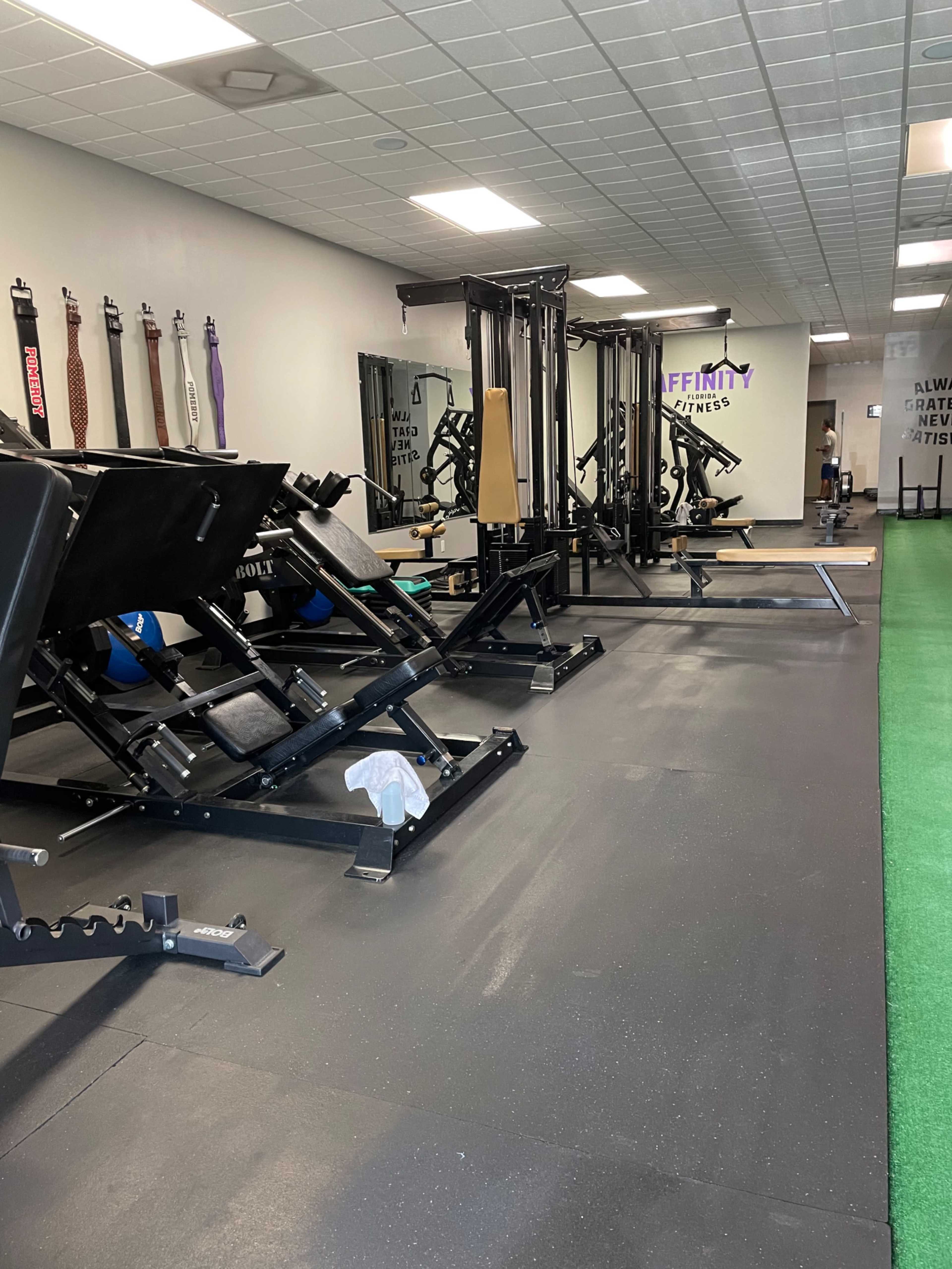 A fitness gym with various exercise machines and equipment arranged in an open space, including a weightlifting area and turf for workouts.