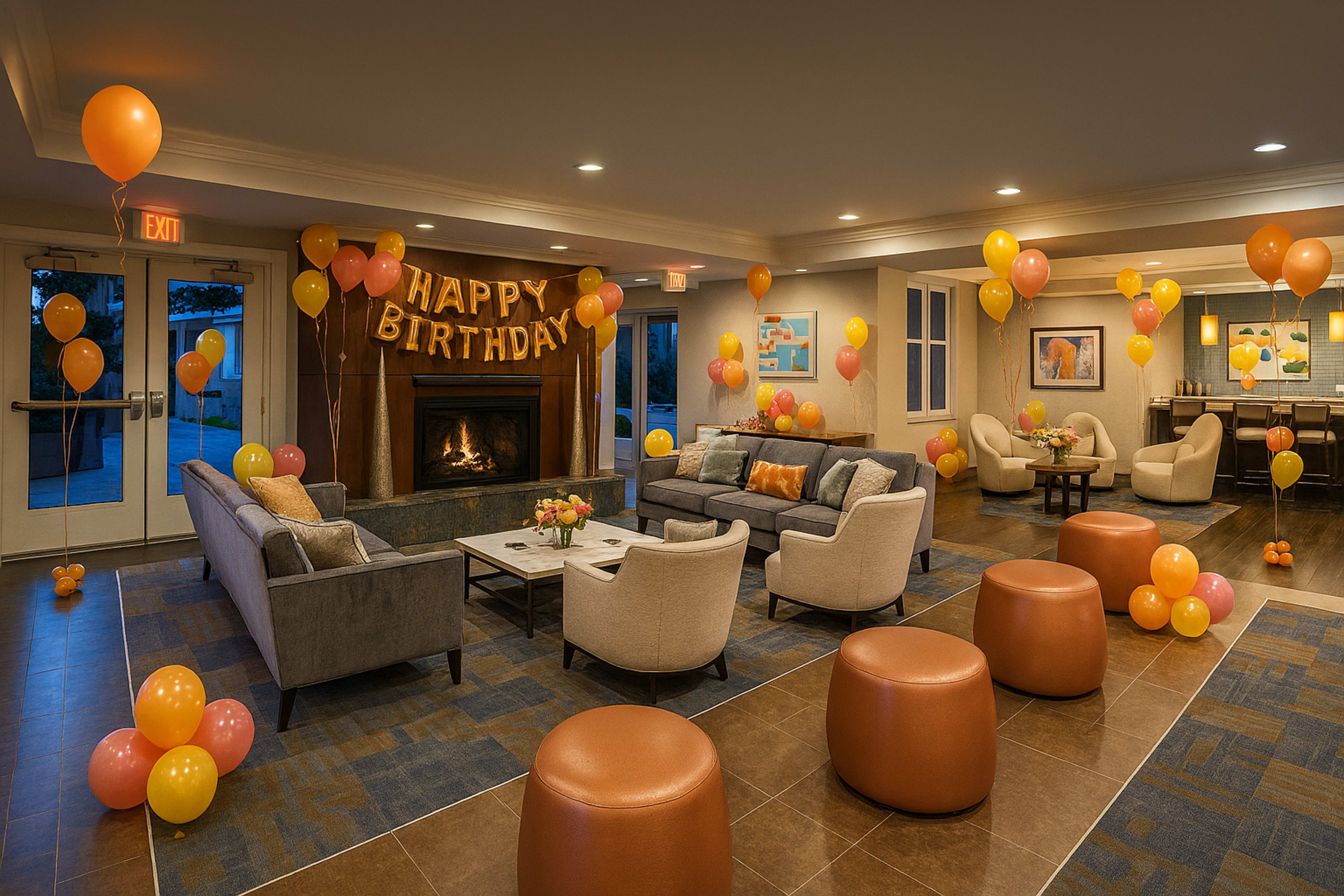 A cozy event space is decorated with balloons and a "Happy Birthday" banner, featuring a fireplace and several seating areas.