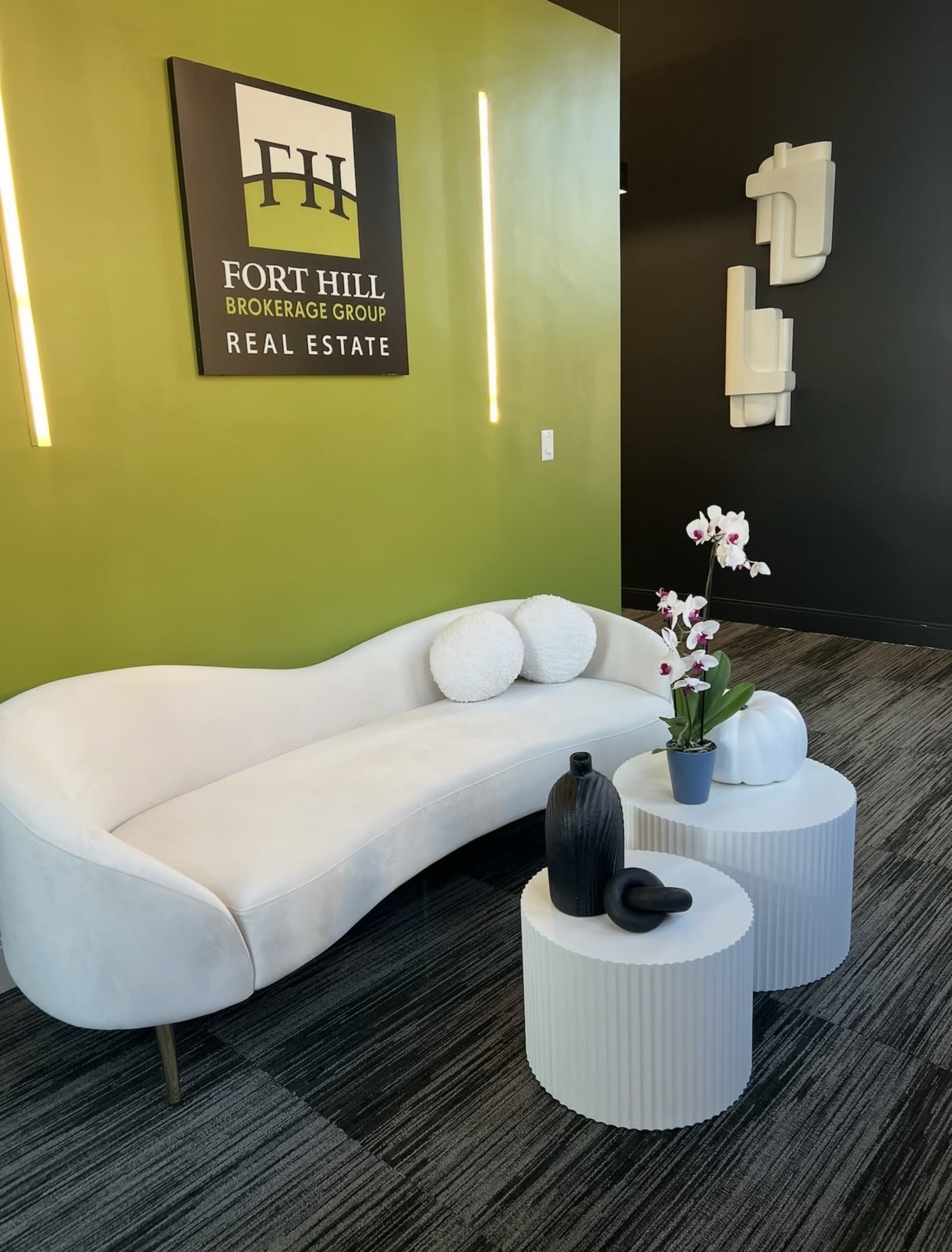 The image shows a modern real estate office reception area featuring a curved white sofa, two round side tables with decorative items, and a green accent wall with the Fort Hill Brokerage Group logo.