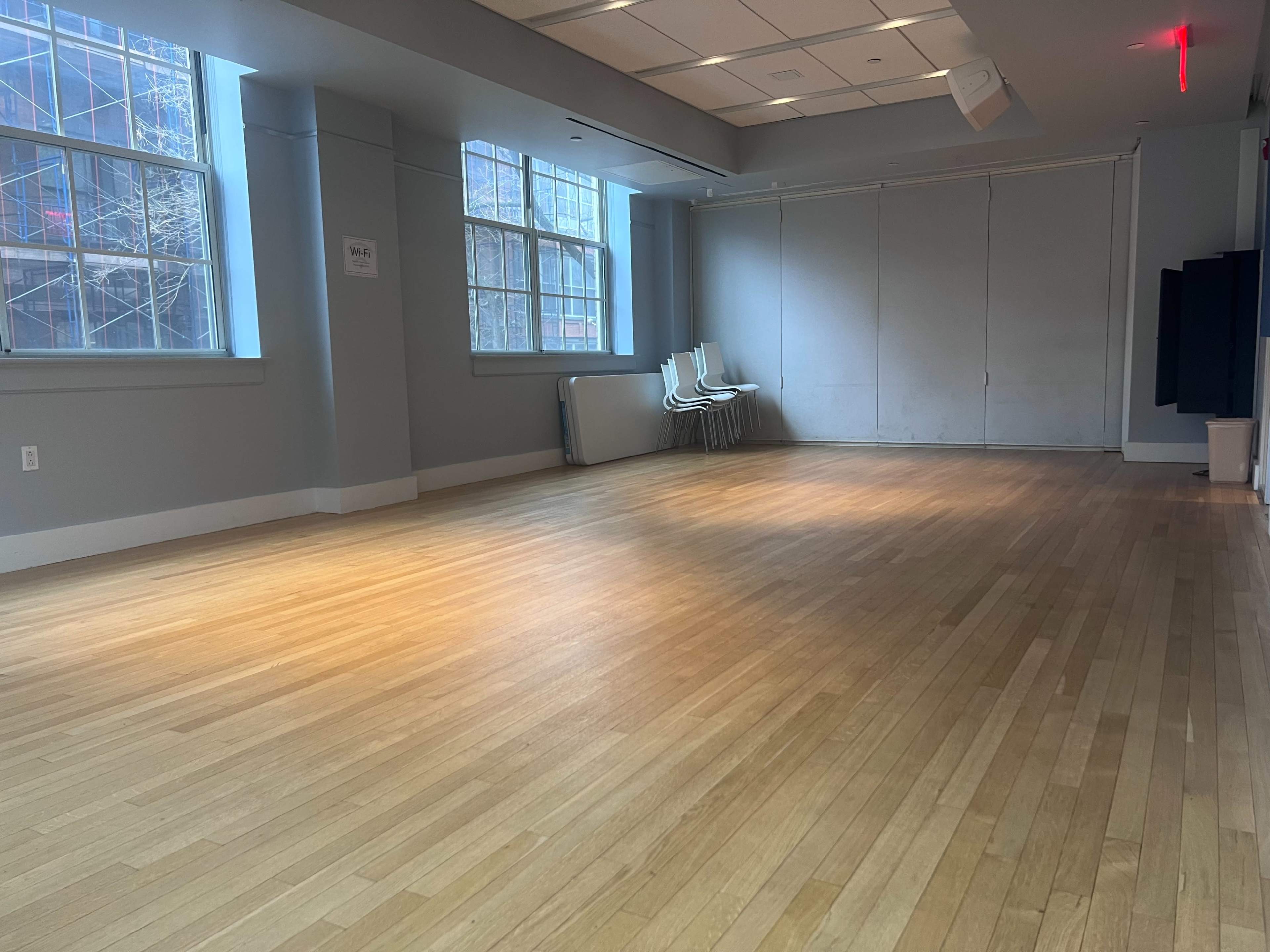 The image shows an empty room with hardwood flooring, large windows letting in natural light, and a stack of chairs against the wall.