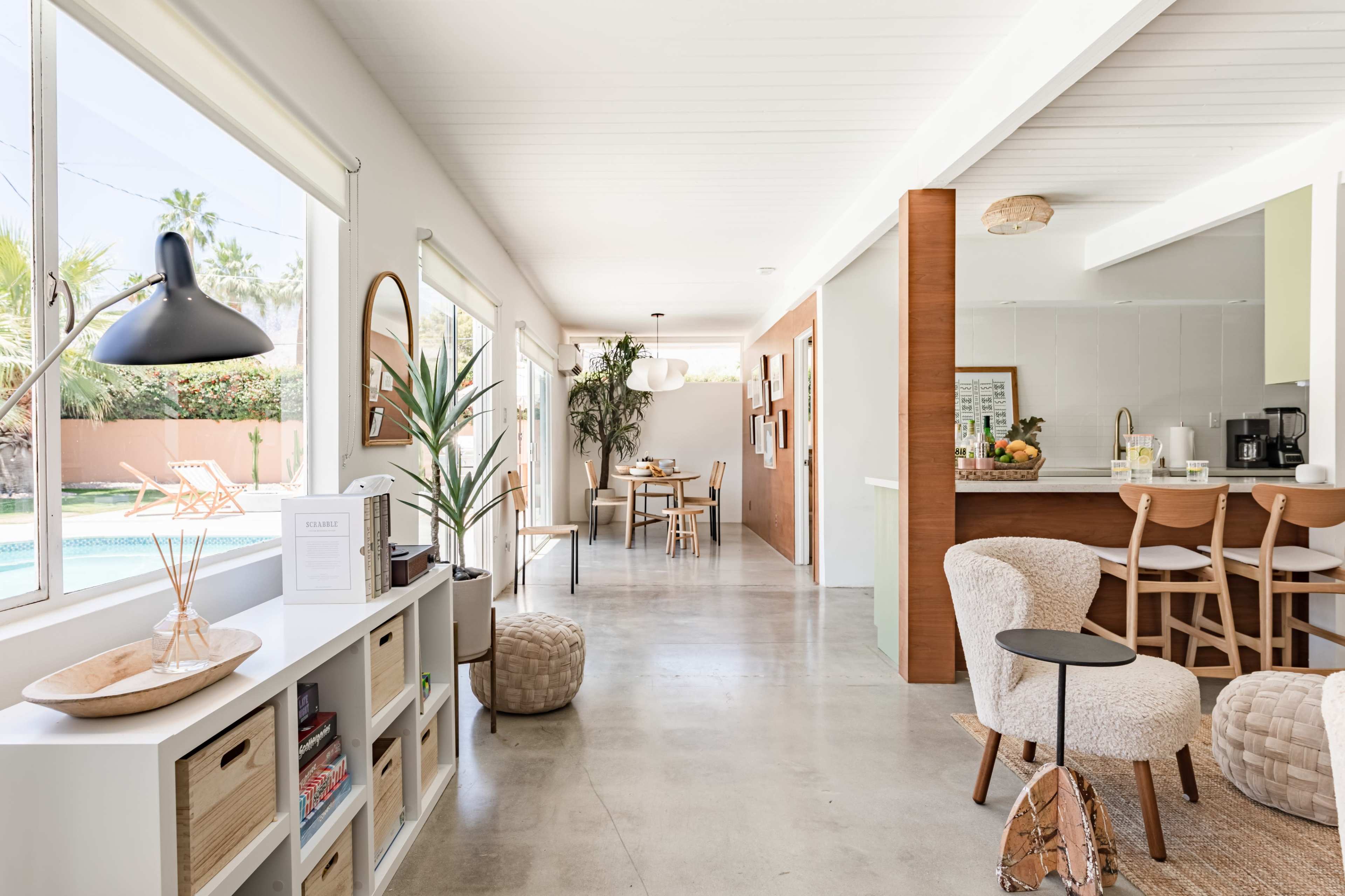 The image shows a modern, open-concept interior featuring a living area with seating arrangements, a dining area in the background, and a view of a pool outside through large windows.