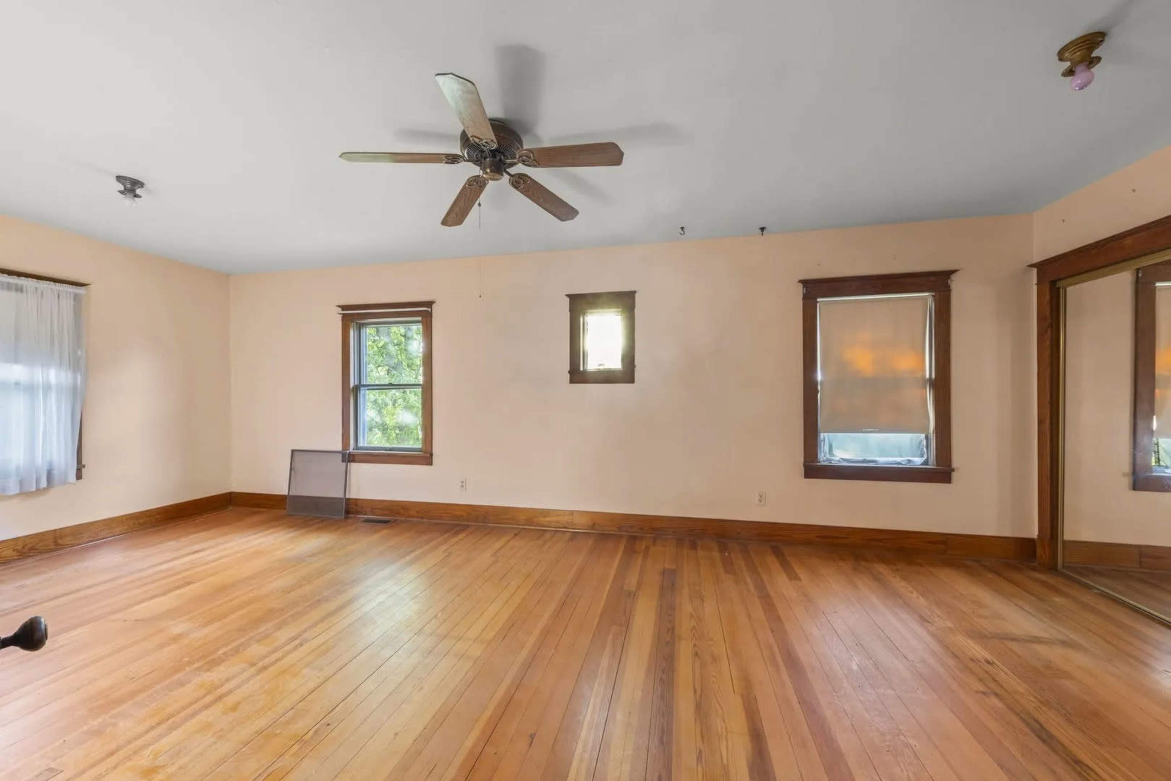 An empty room with wooden flooring, a ceiling fan, and two windows, featuring light-colored walls and a mirrored closet.