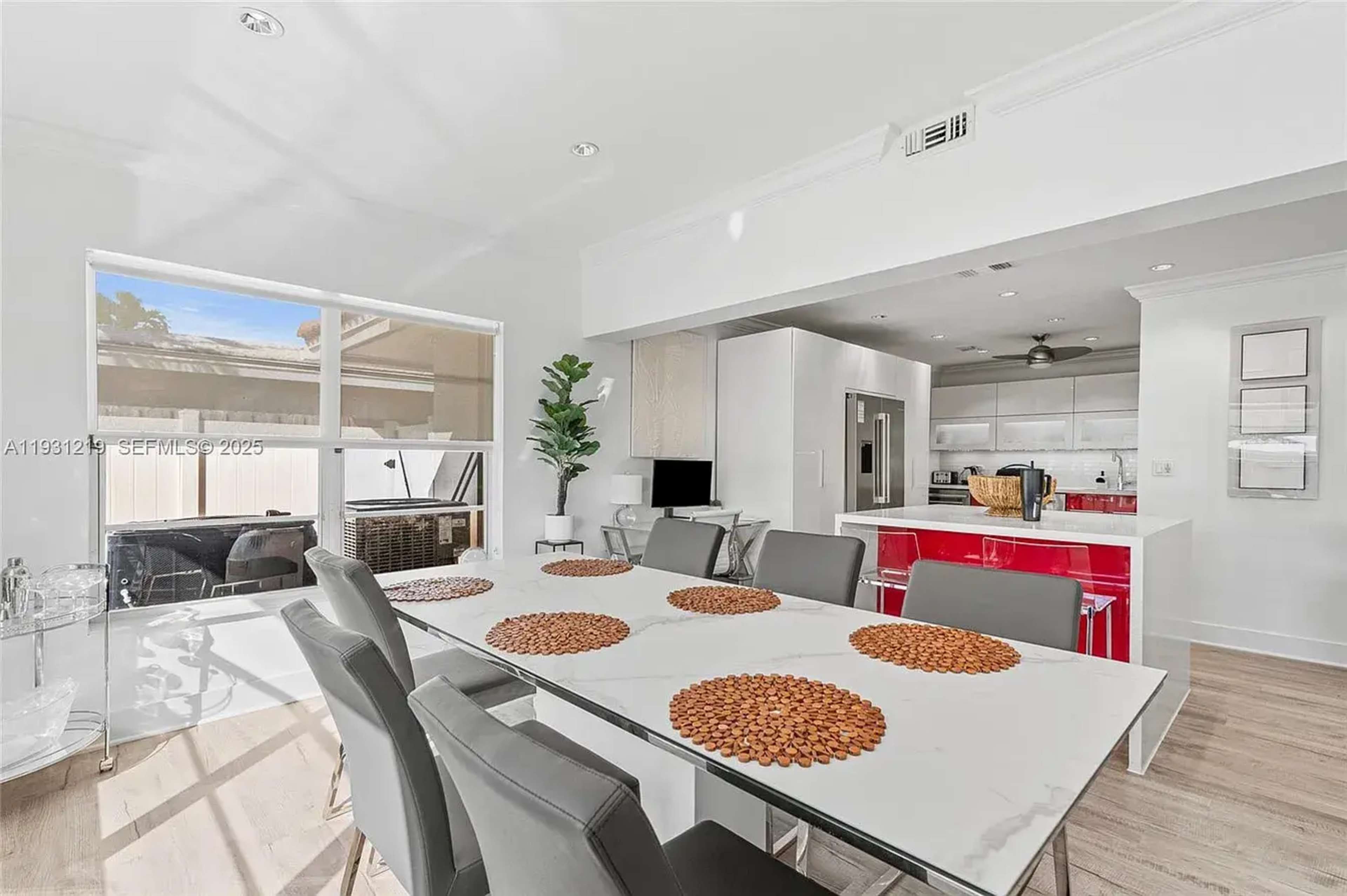 A modern dining area features a glass table with decorative placemats and sleek chairs, adjacent to an open kitchen with white cabinetry and red accents.