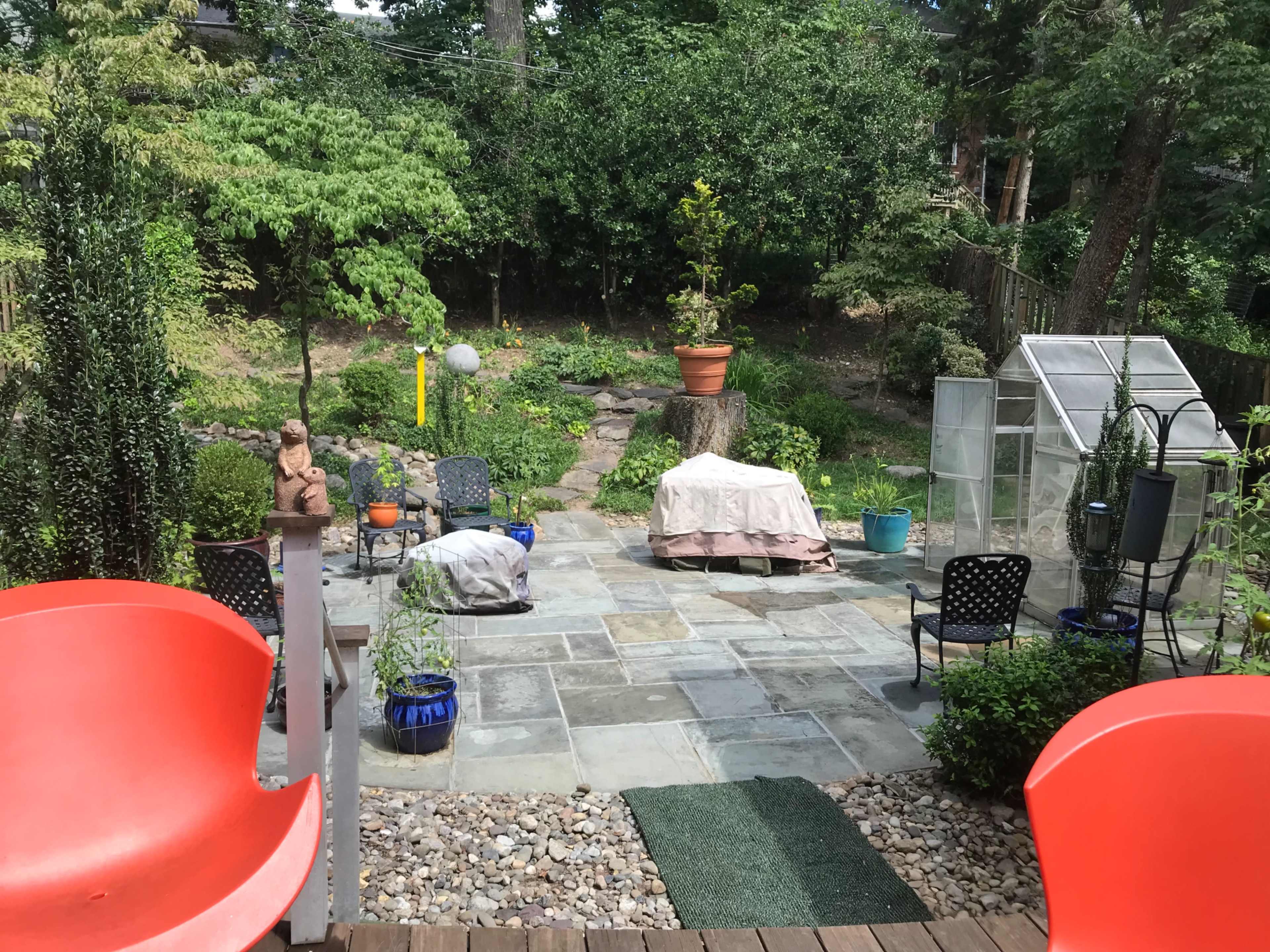A backyard patio with stone flooring, surrounded by greenery, featuring several chairs, a potted plant, and a greenhouse in the background.