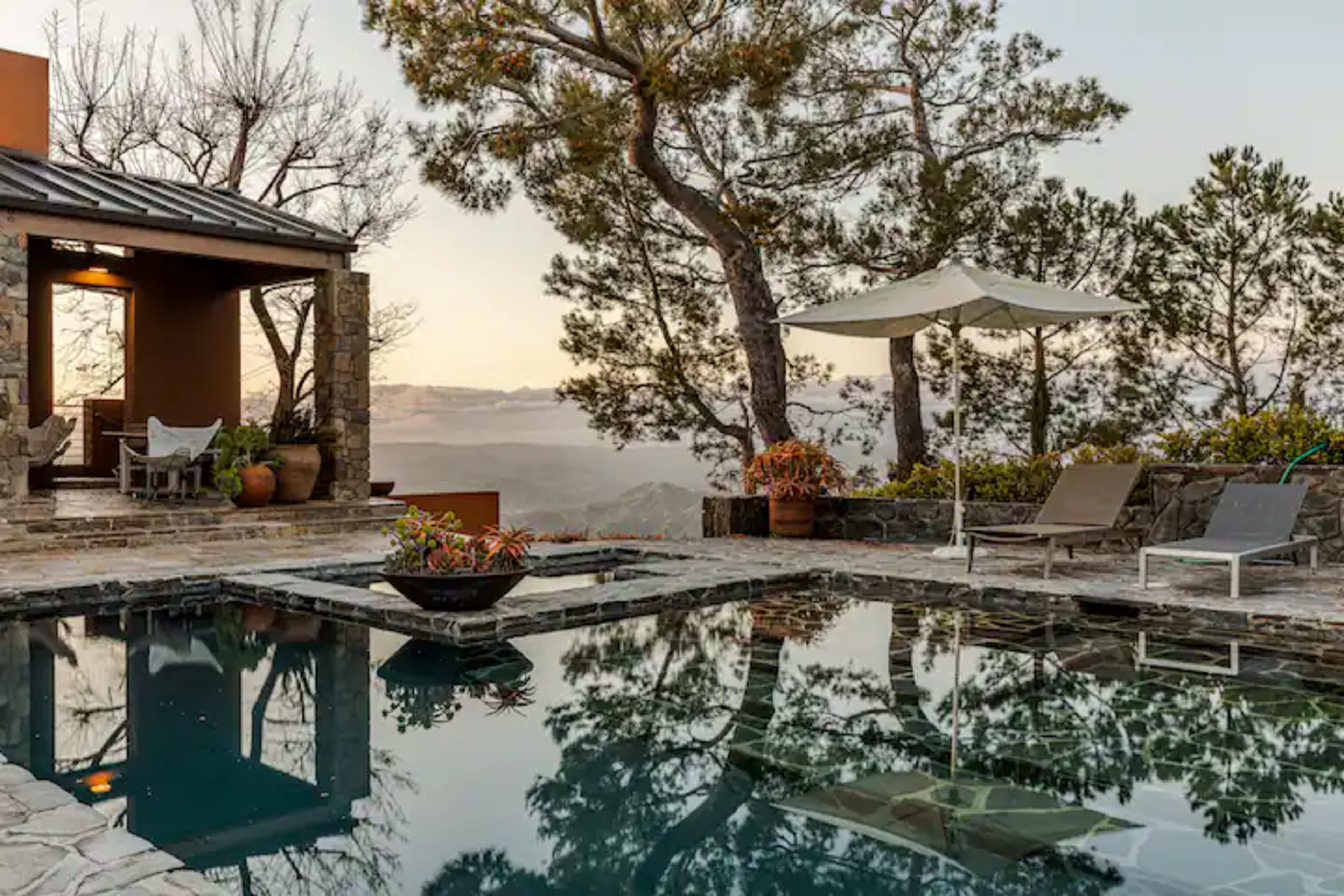 The image shows a modern stone pool area surrounded by trees, with a patio featuring seating and an umbrella.