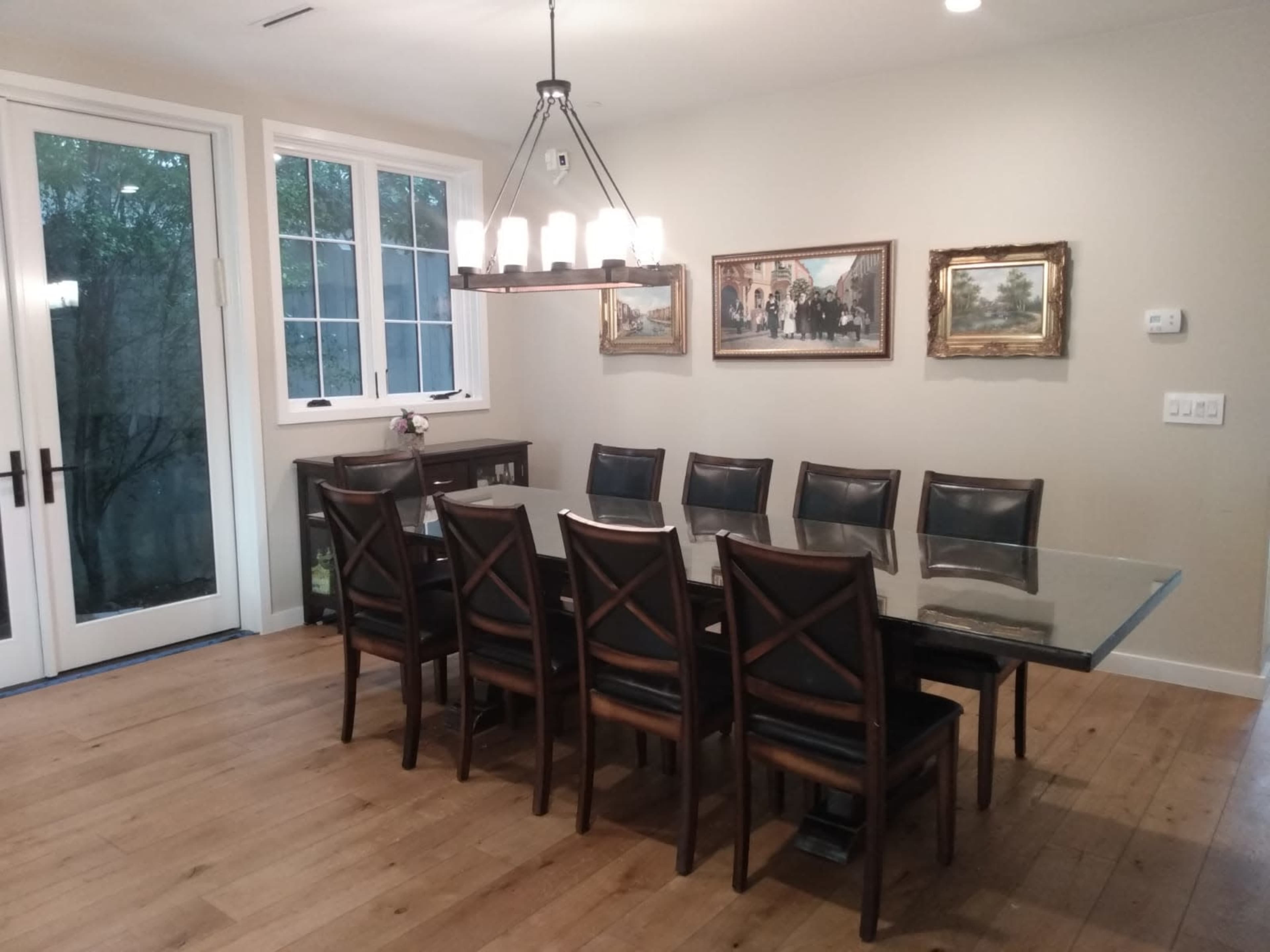 A dining room featuring a long glass table with eight black leather chairs, and a chandelier overhead, along with two paintings on the wall.