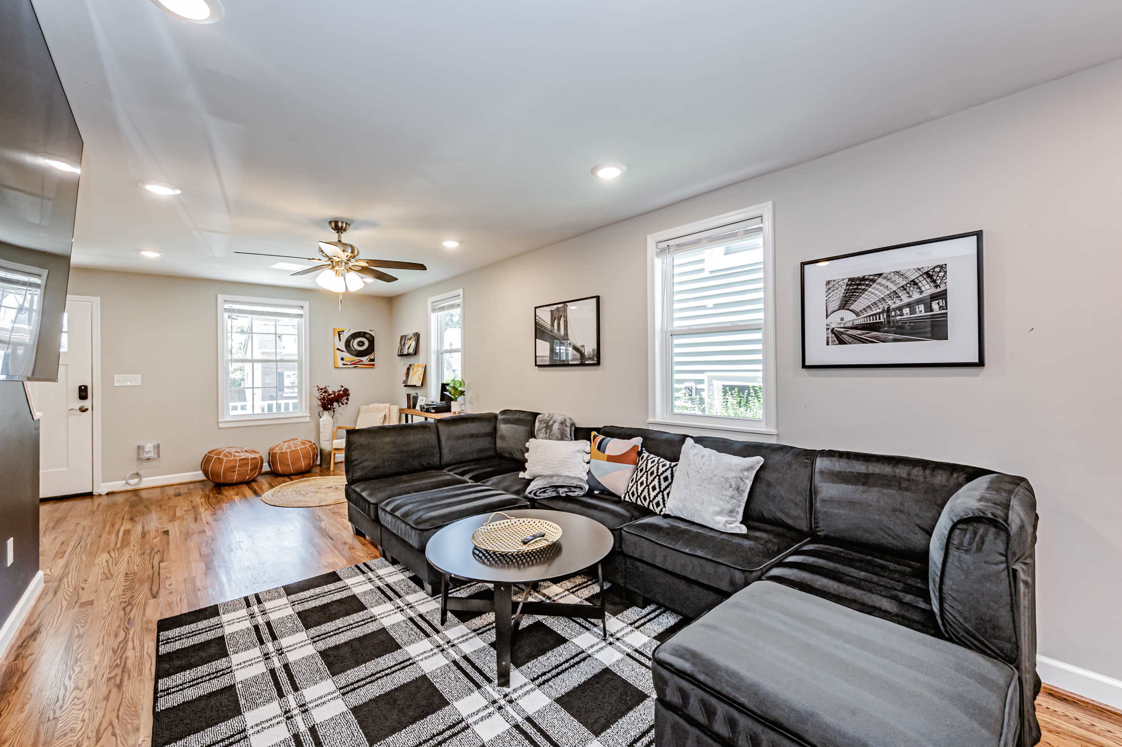 The image shows a modern living room with a sectional sofa, a coffee table, and two windows that provide natural light, along with a decorative rug on the hardwood floor.