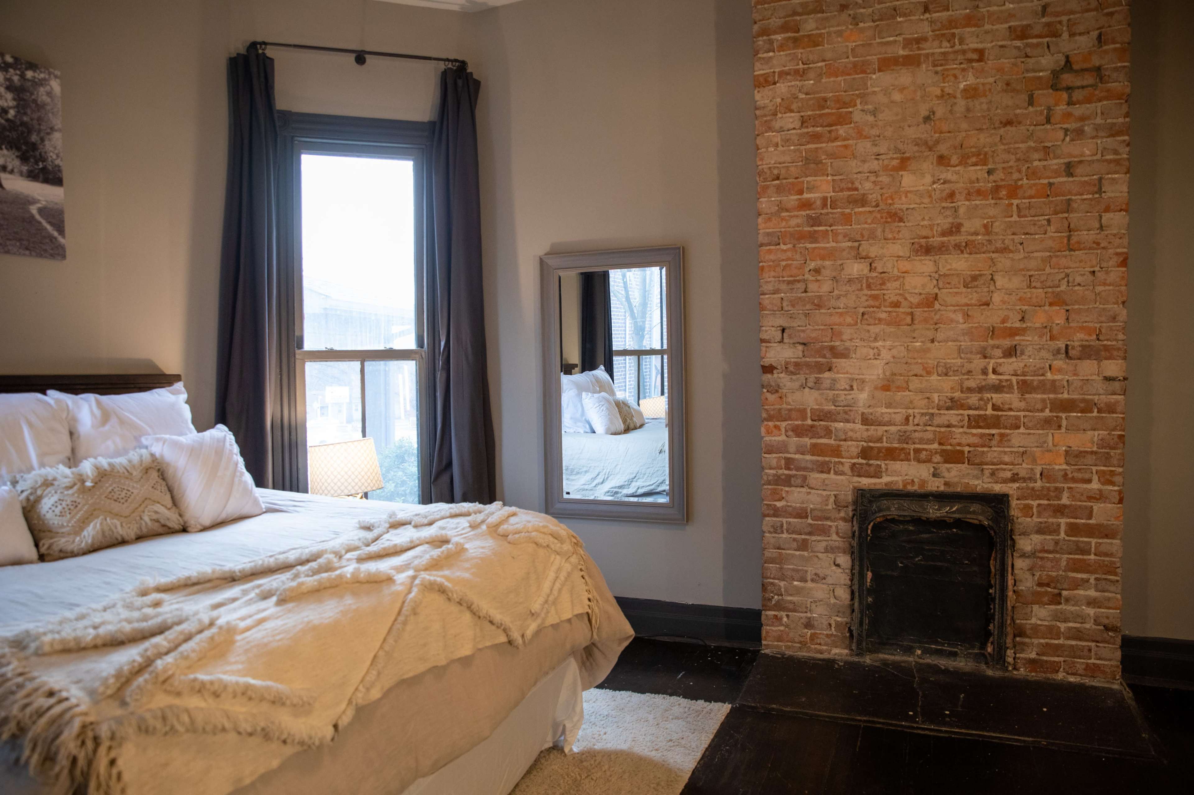 A bedroom featuring a bed with a textured blanket, a brick fireplace, and a large window with grey curtains.