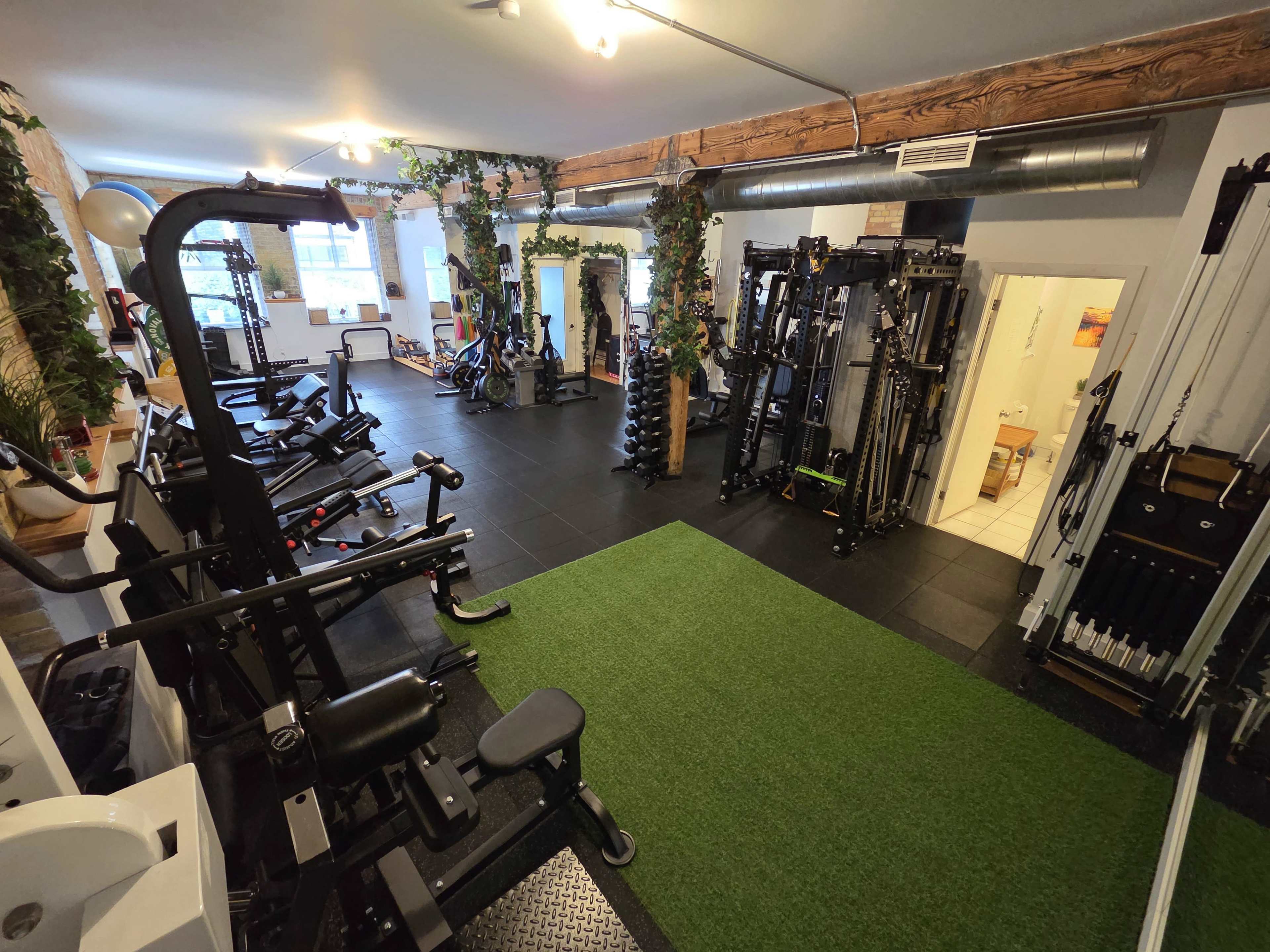 The image shows a spacious gym area equipped with various exercise machines, free weights, and green turf flooring, complemented by plants and bright lighting.