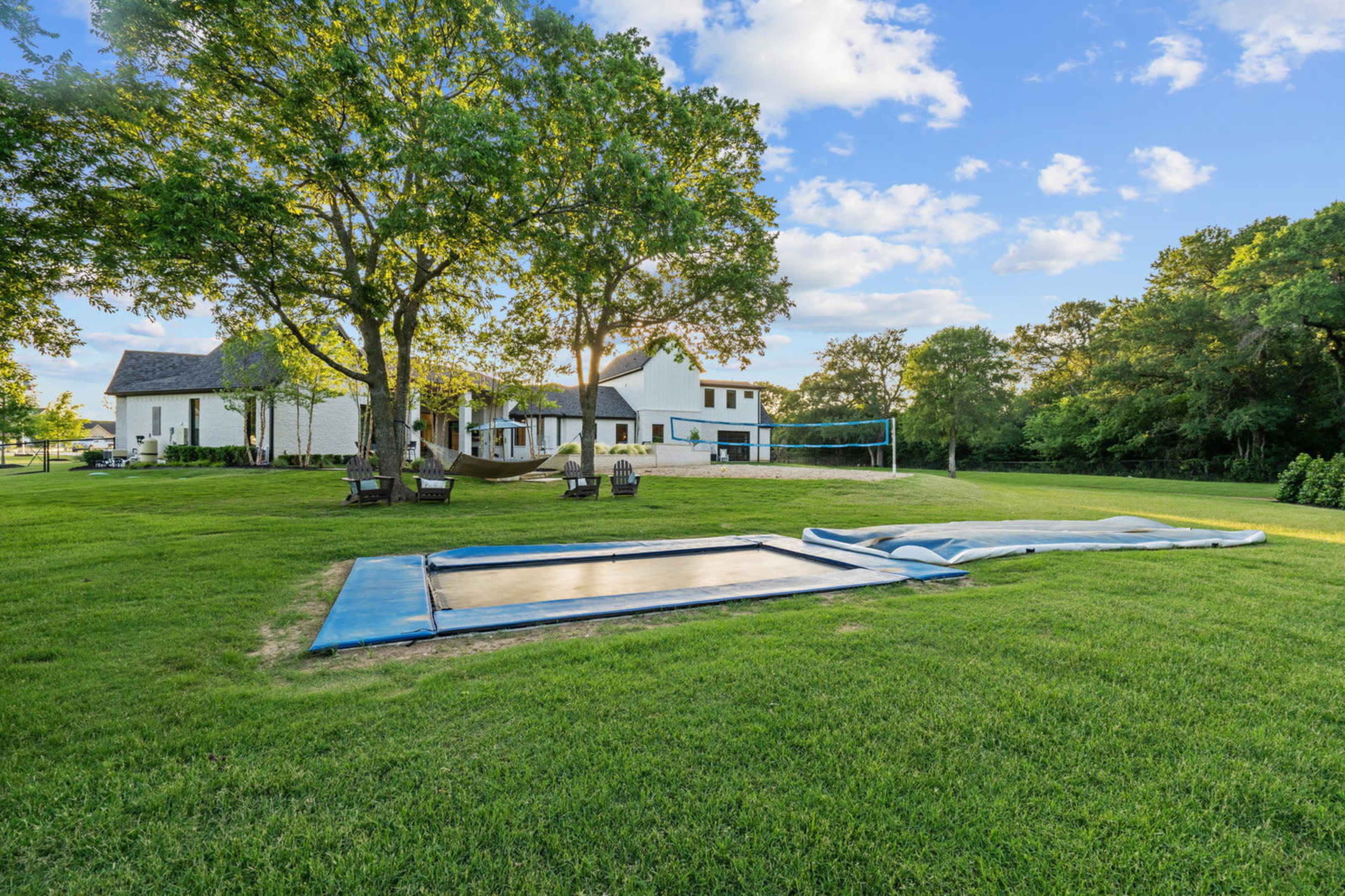 A grassy yard featuring a trampoline and a volleyball net beside a white house and trees in the background.