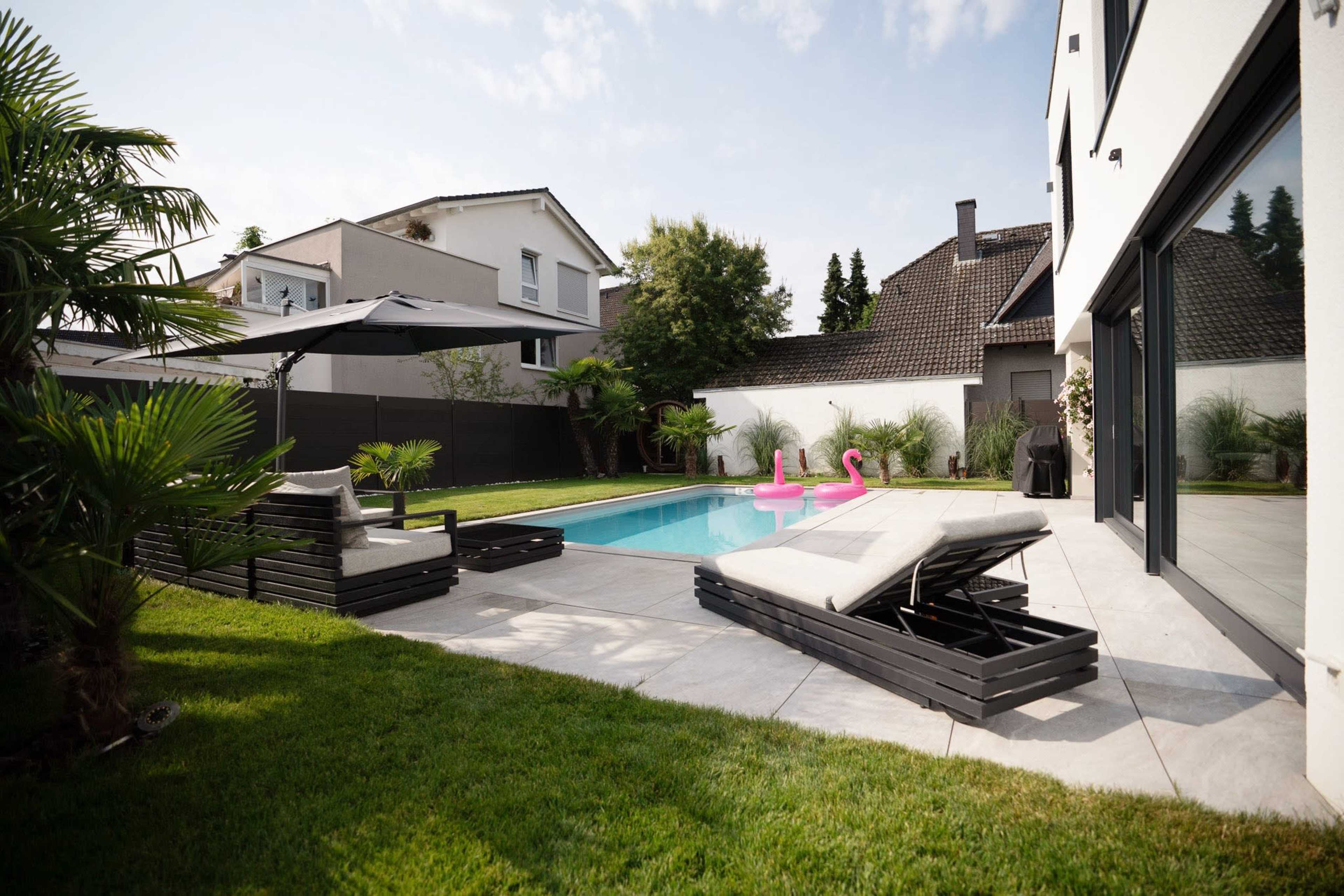 The image depicts a residential backyard with a swimming pool, two pink flamingo floaties, a lounge chair, and a shaded seating area surrounded by greenery.