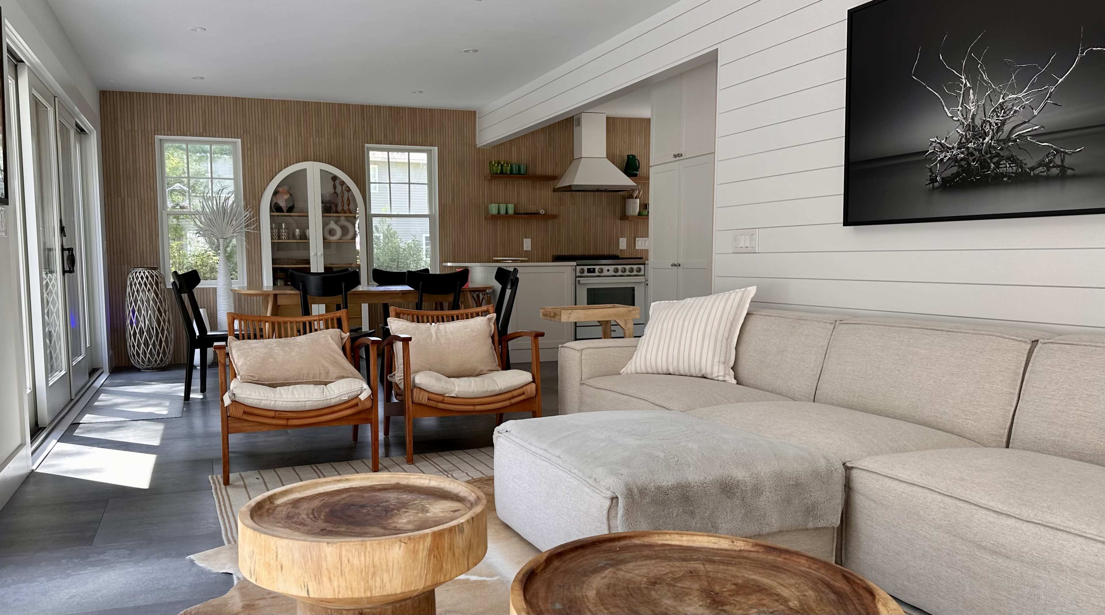 A modern living space with a beige sectional sofa, wooden accent chairs, round coffee tables, and a dining area in the background.