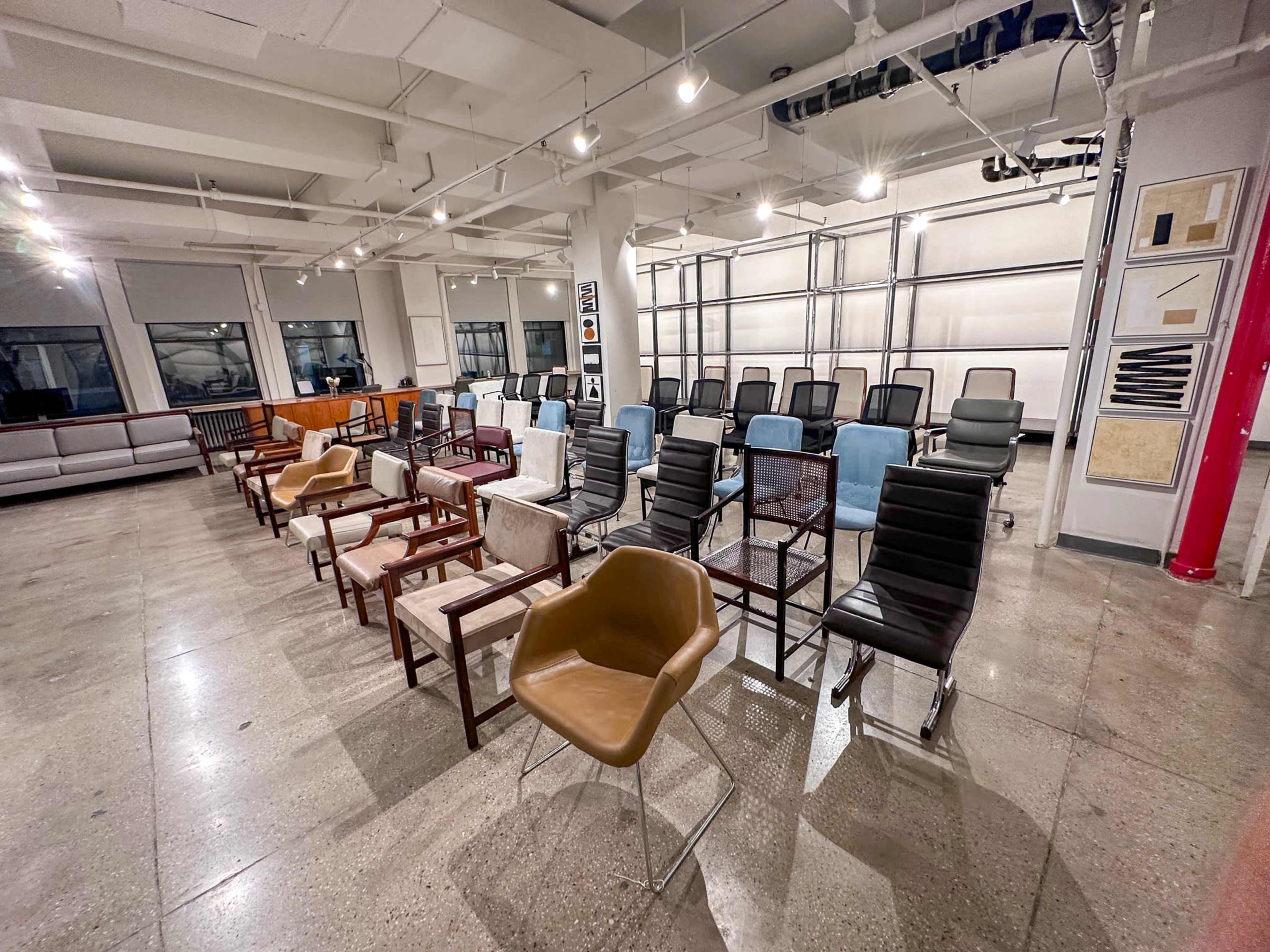 The image shows a spacious room arranged with various types of chairs set up in rows, with large windows and overhead lighting.