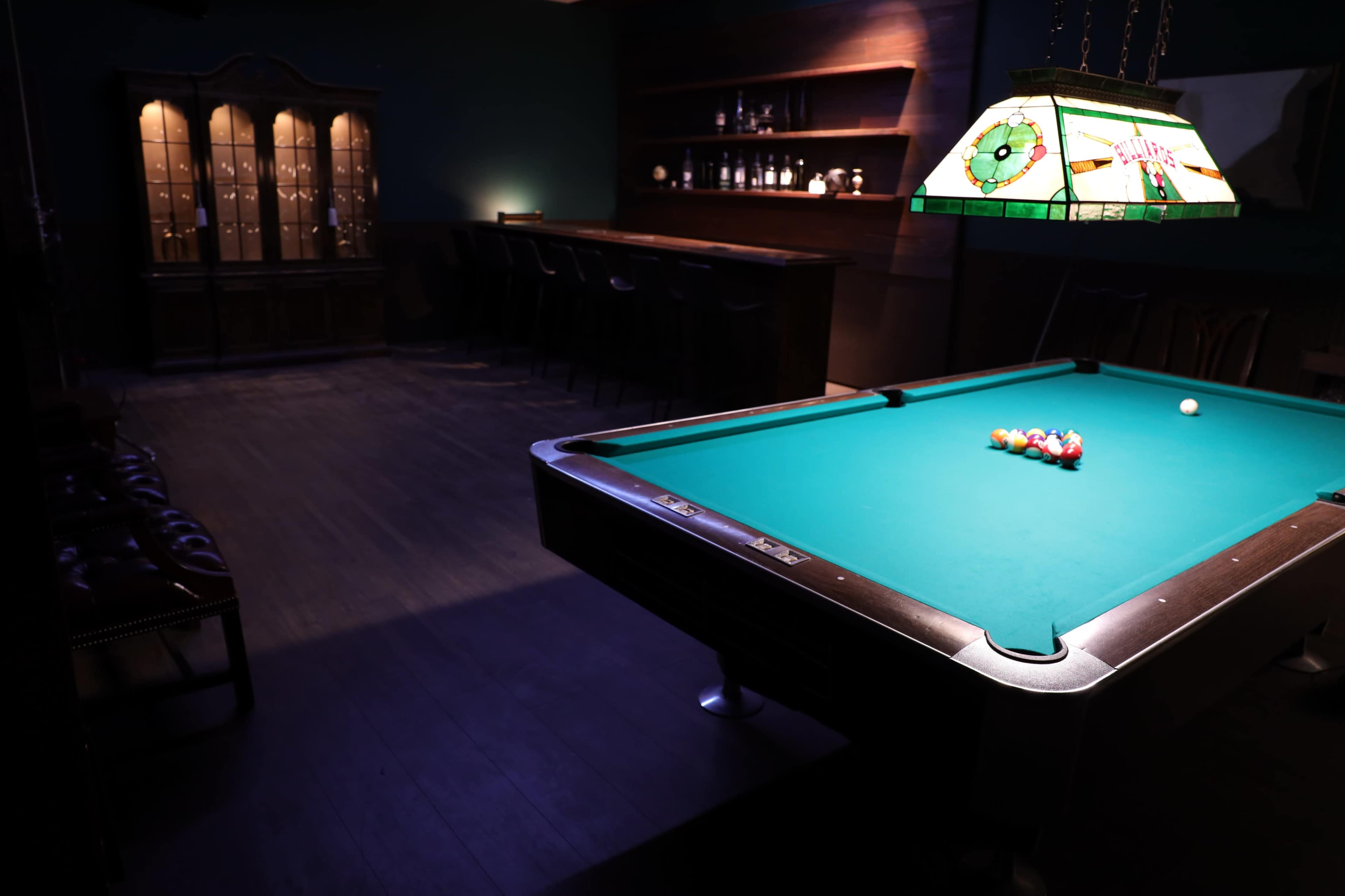 The image shows a dimly lit room featuring a billiards table with colorful balls, a bar area in the background, and a glass cabinet displaying items.