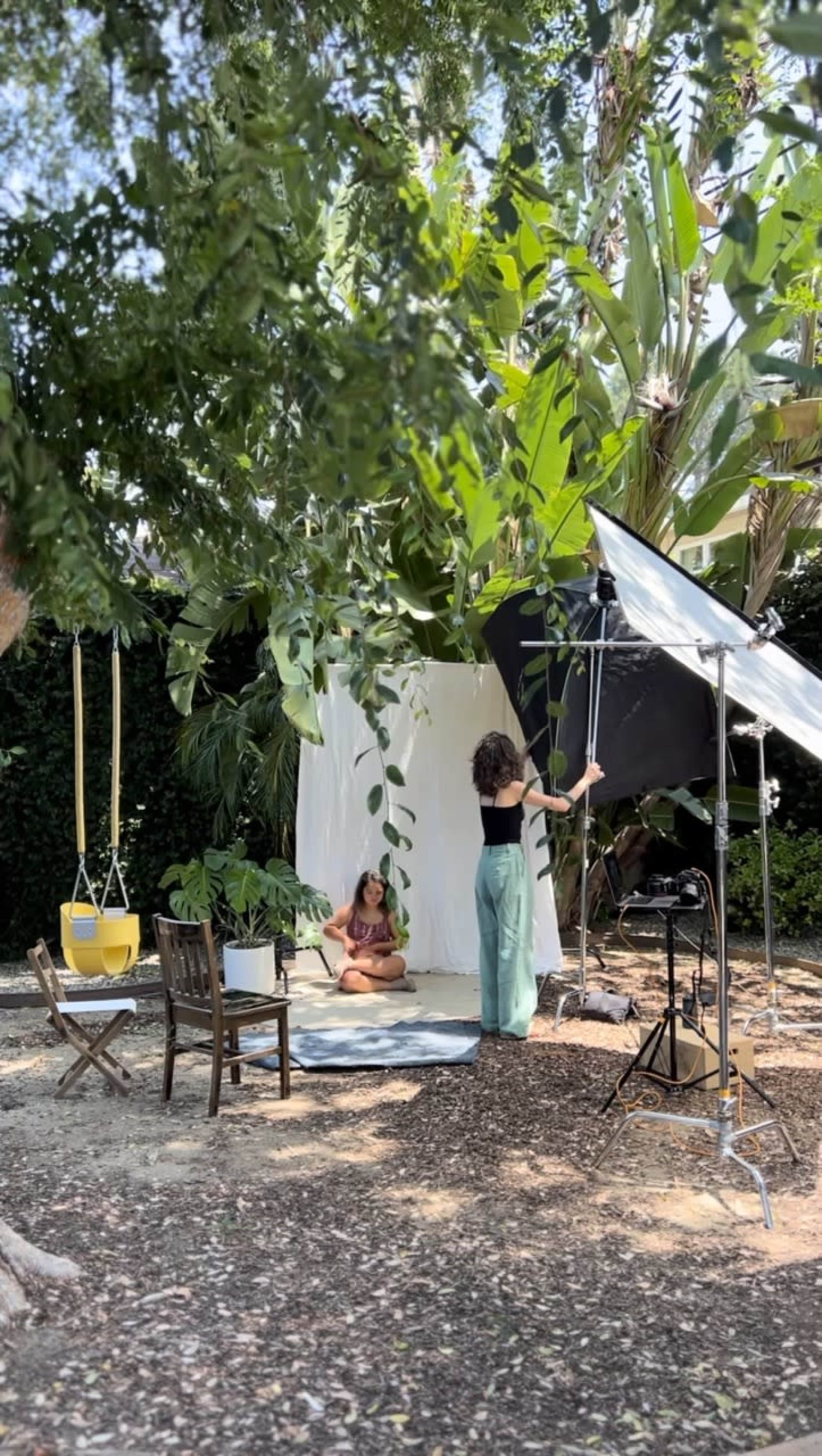 A woman adjusts lighting equipment while another sits on a mat in a garden setting surrounded by tropical plants.
