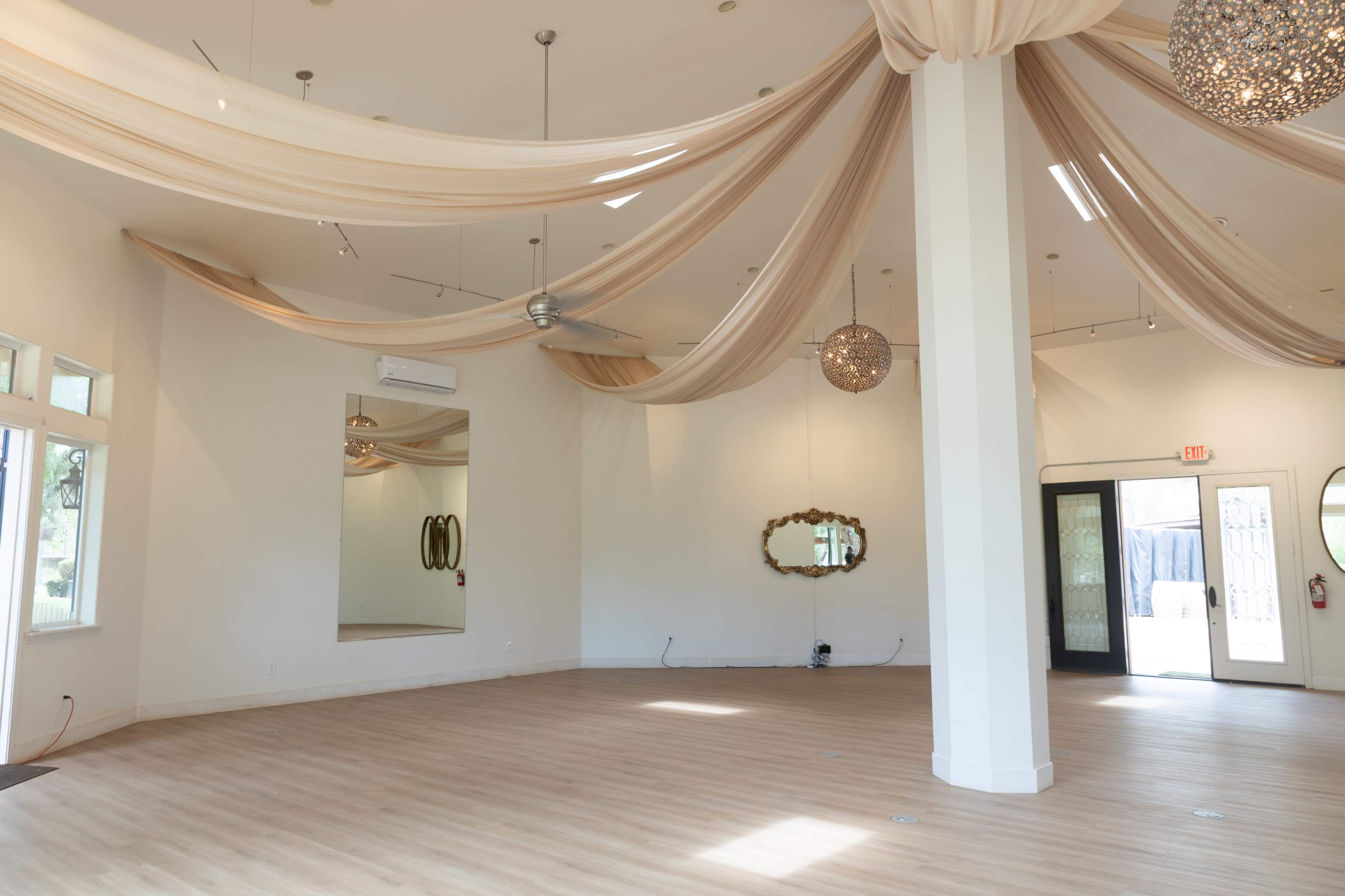 The image shows an empty interior space with draped fabric overhead, a large mirror on one wall, and decorative light fixtures.