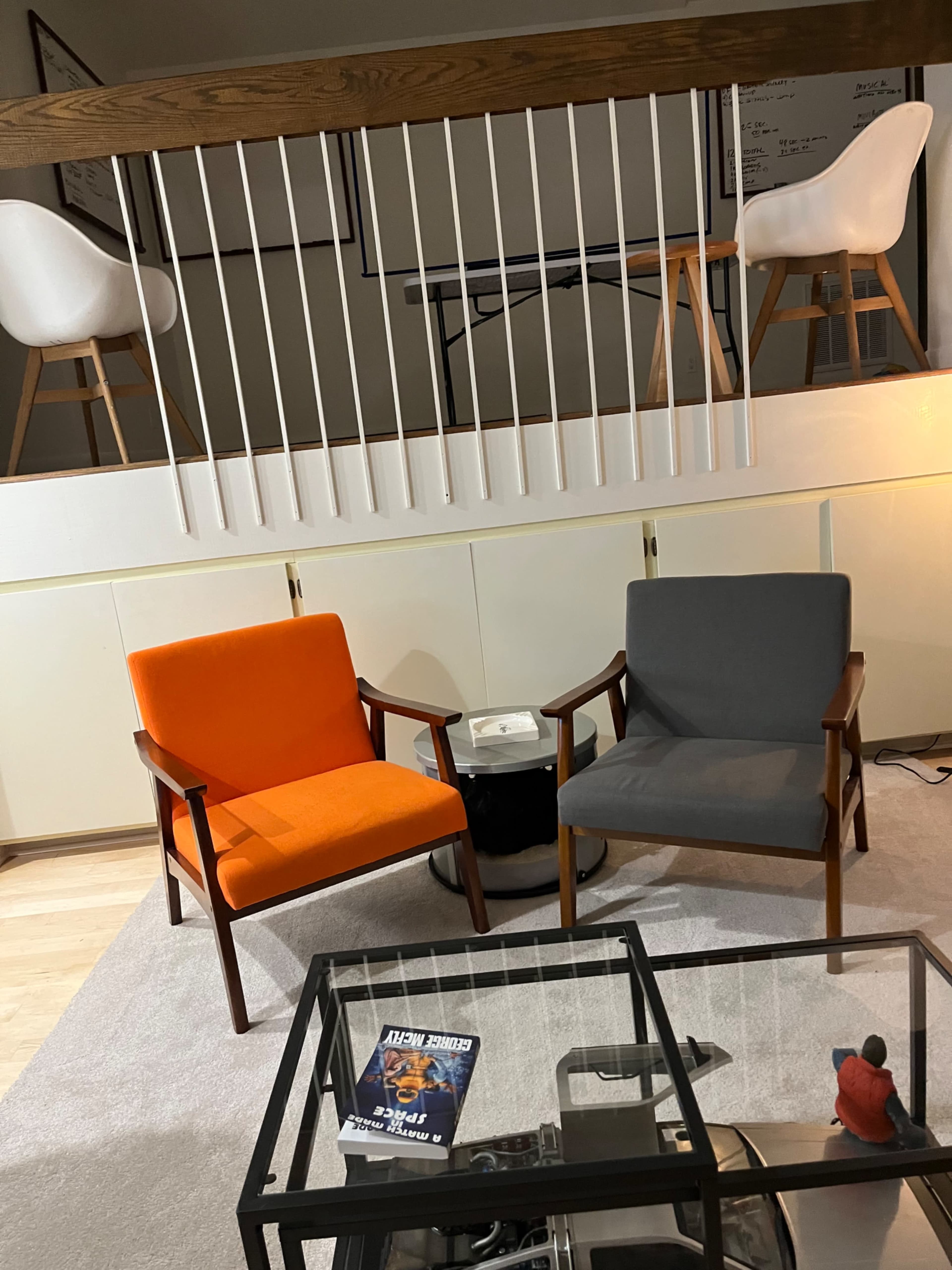 The image shows a modern living space with two armchairs—a bright orange one and a gray one—situated around a glass coffee table on a light-colored rug.