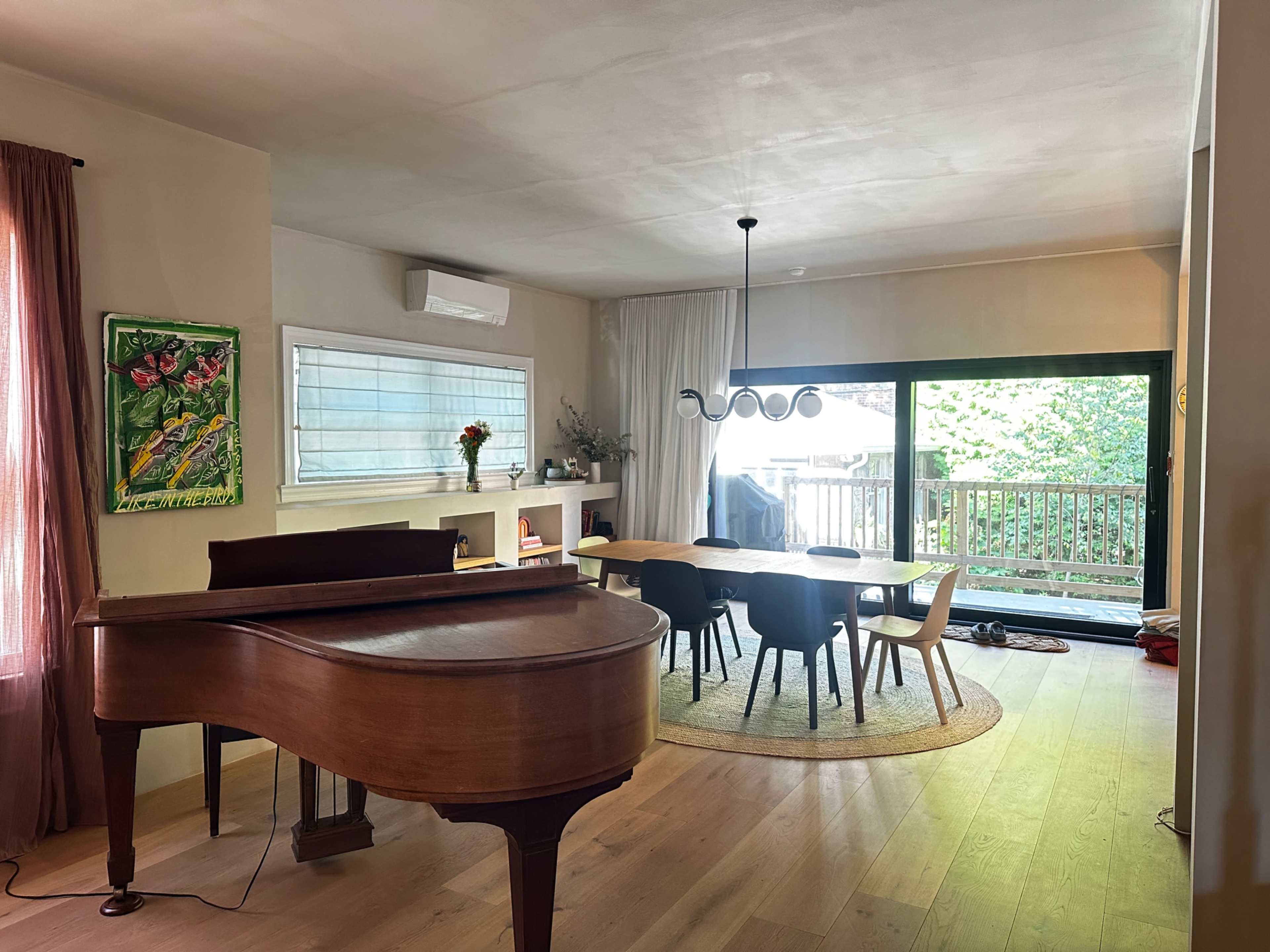 The image shows a modern living space featuring a grand piano in the foreground and a dining area with a round table surrounded by chairs, illuminated by natural light from large windows.