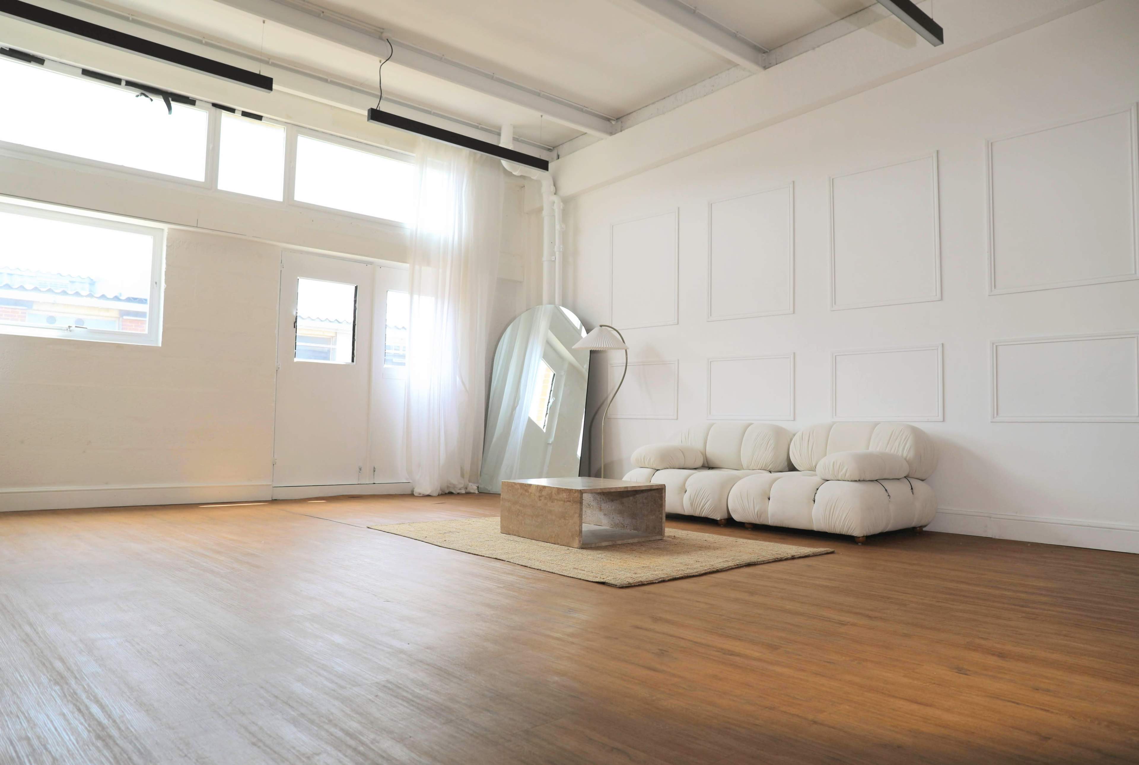 A minimalistic room with a white sofa, a large mirror, and a wooden floor, featuring ample natural light from several windows.