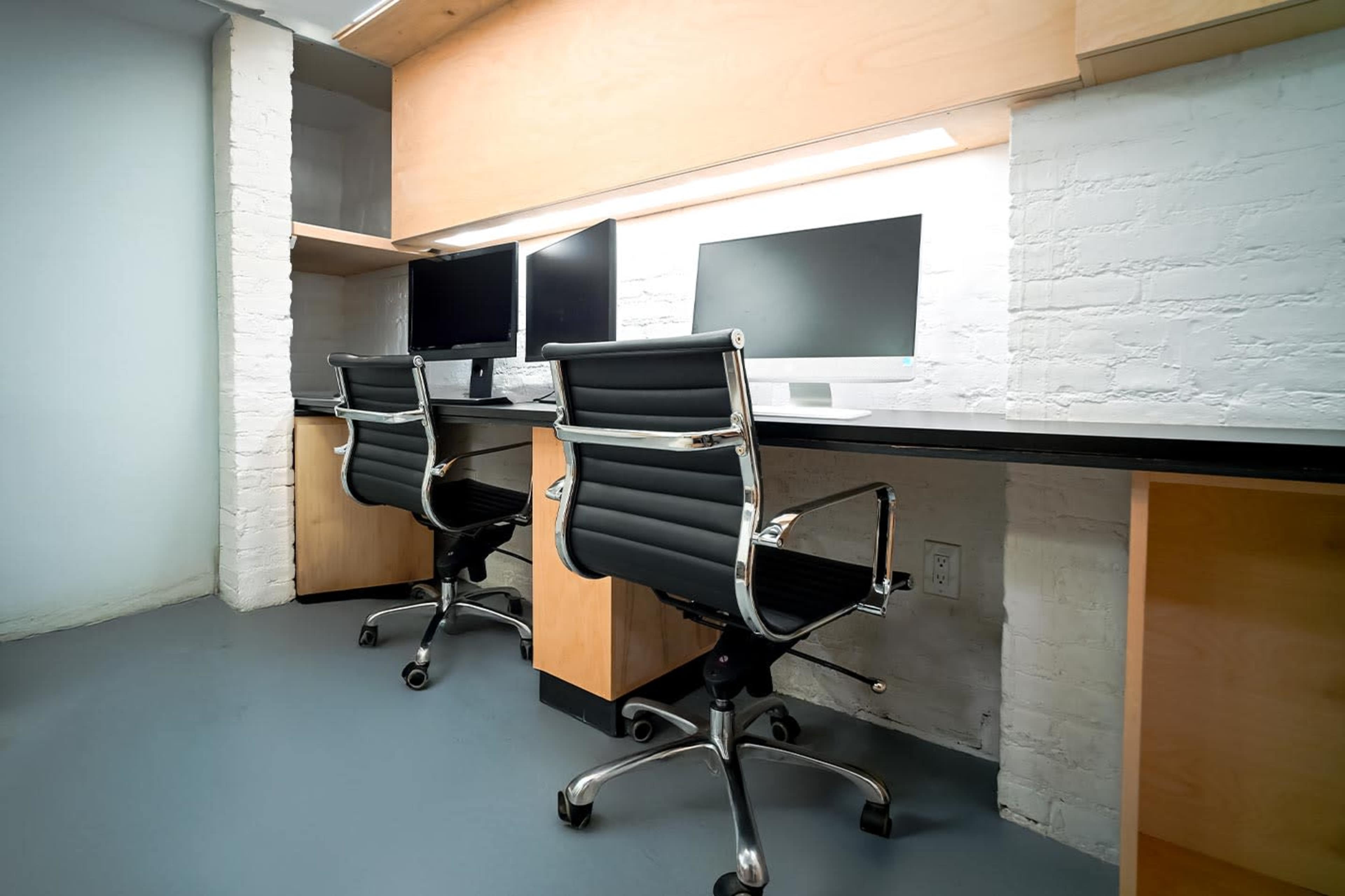 The image shows an office space featuring two modern workstations with black swivel chairs and dual computer monitors on each desk, placed against a brick wall.