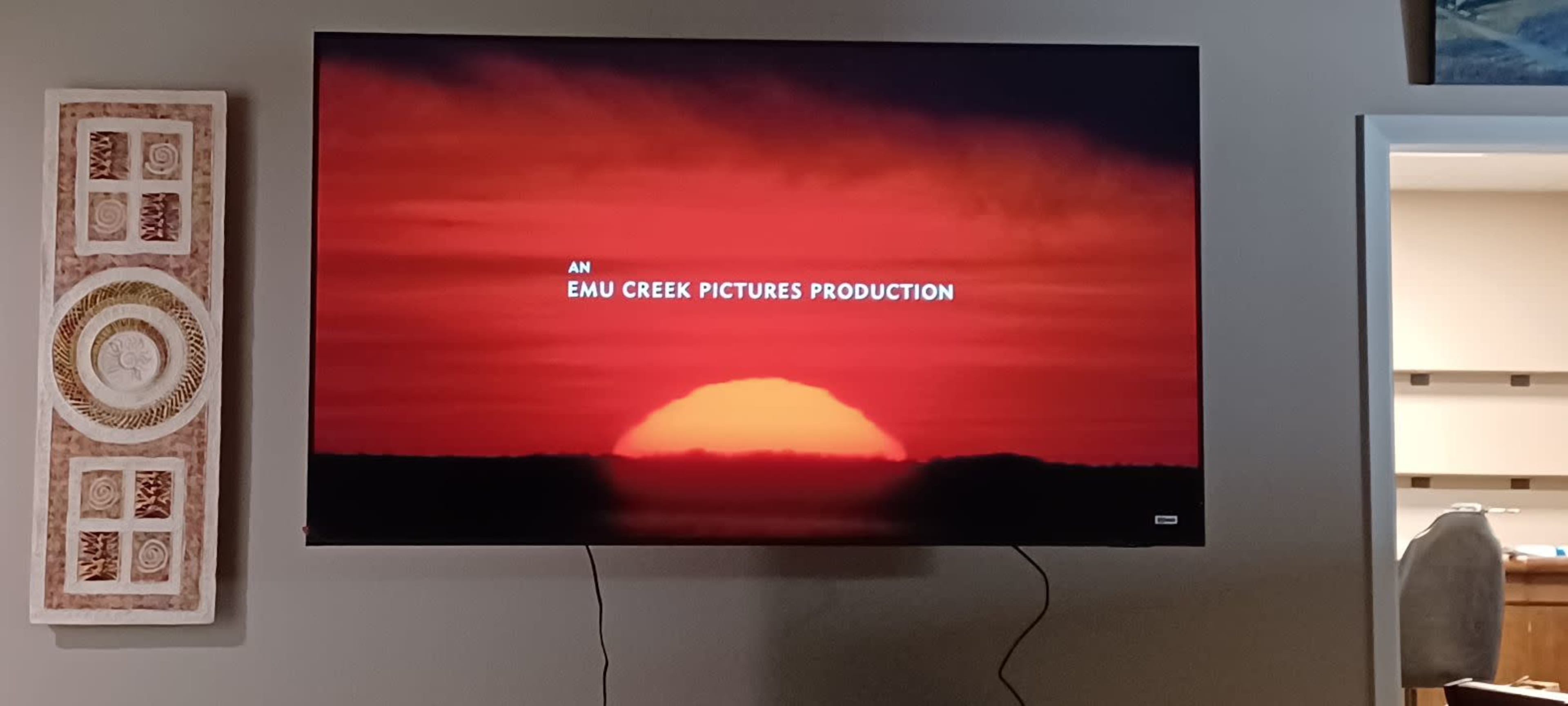 The image shows a television displaying the text "AN EMU CREEK PICTURES PRODUCTION" with a sunset backdrop.