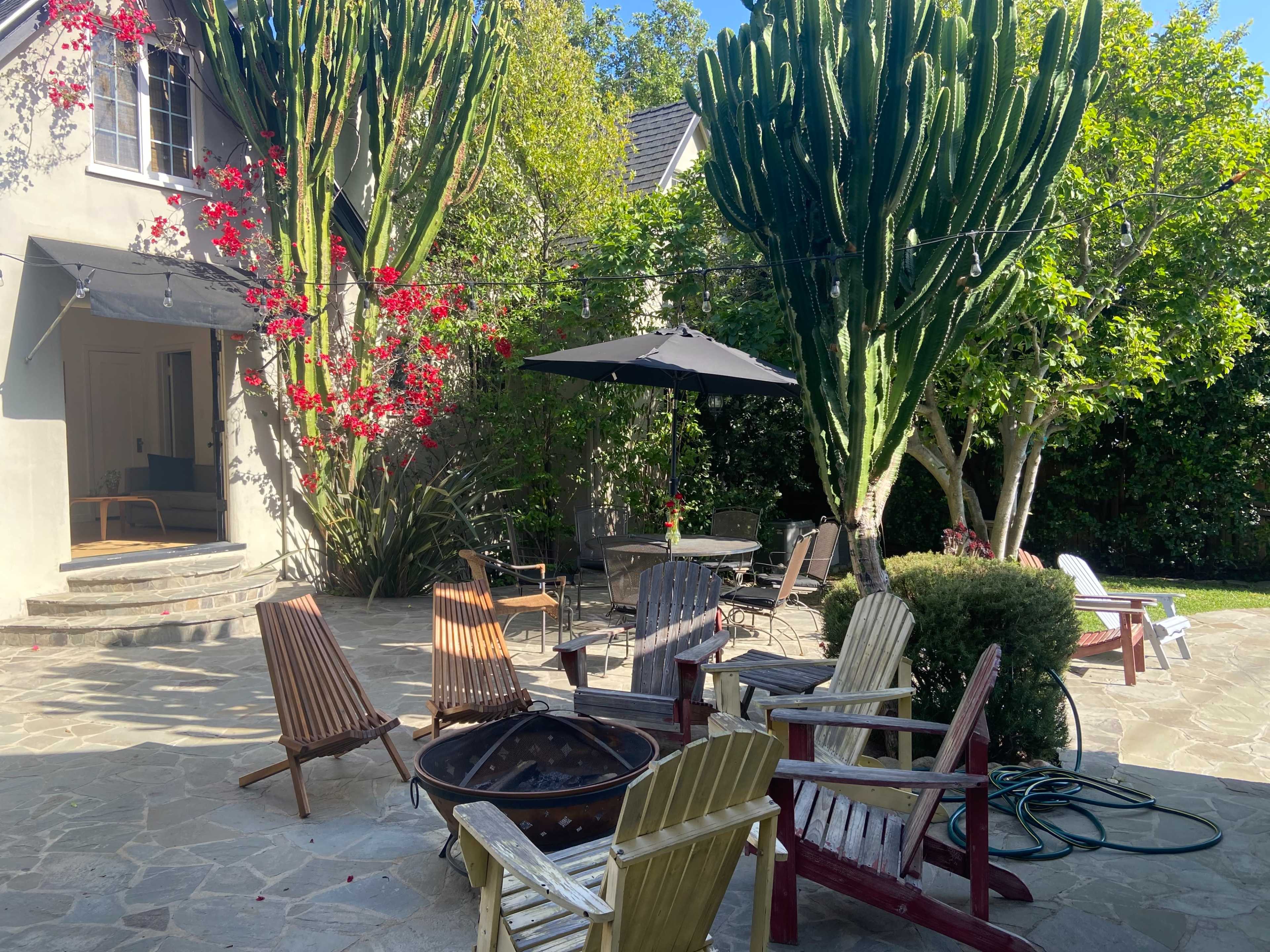 A backyard patio features several wooden chairs arranged around a fire pit, with a large umbrella and tall cacti in the background.