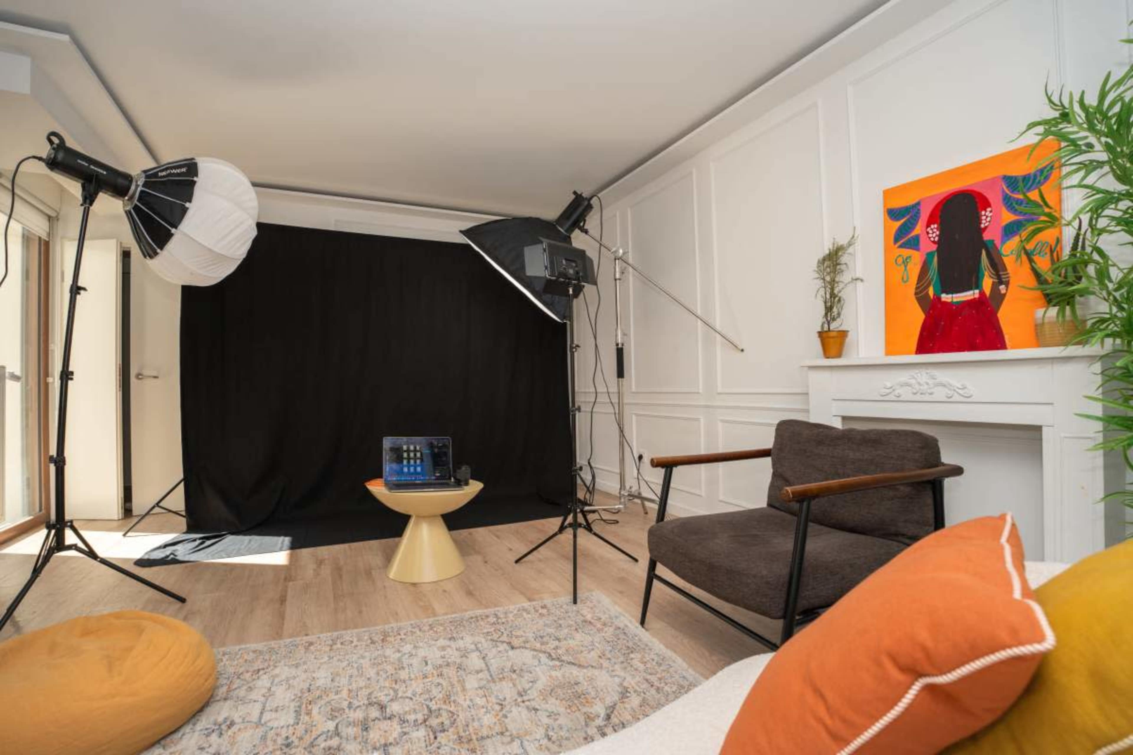 The image shows a photography studio setup featuring a black backdrop, two lighting fixtures, a small round table with equipment, and a colorful artwork on the wall.