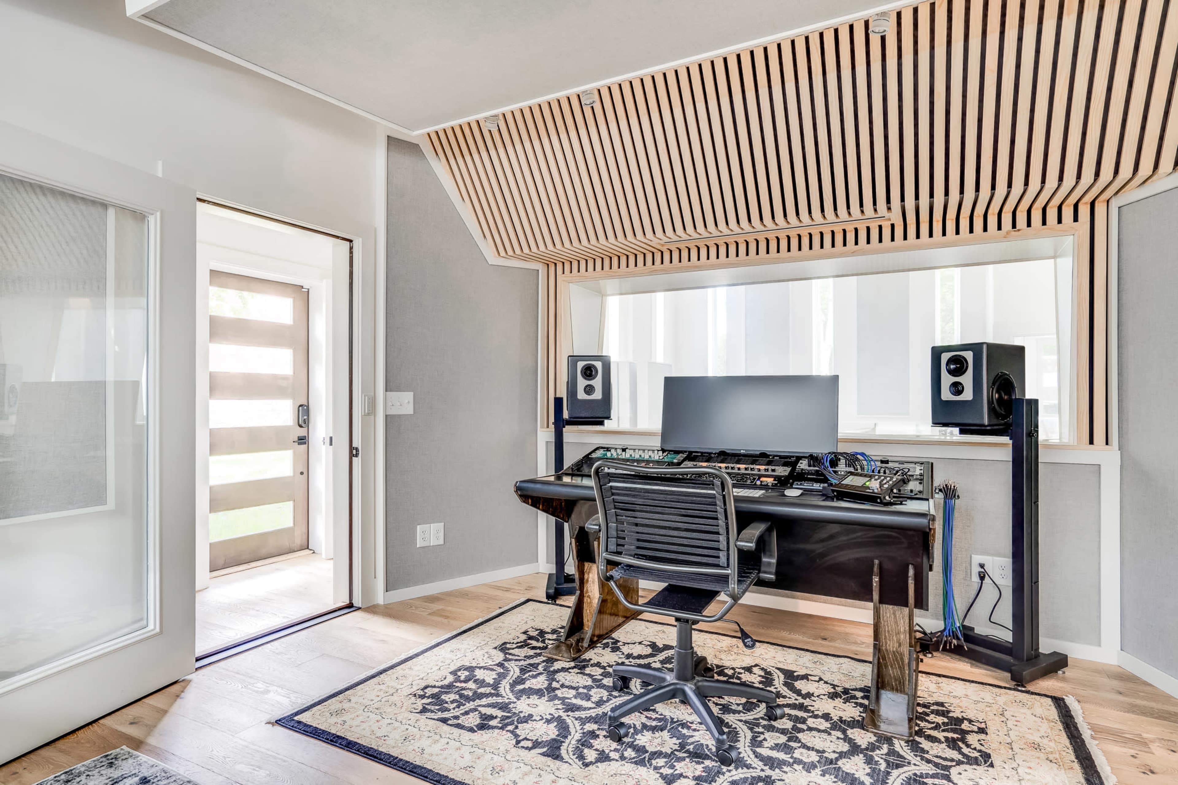 A modern recording studio with a desk, computer monitor, and sound equipment, featuring a wooden slatted ceiling and a glass door leading outside.