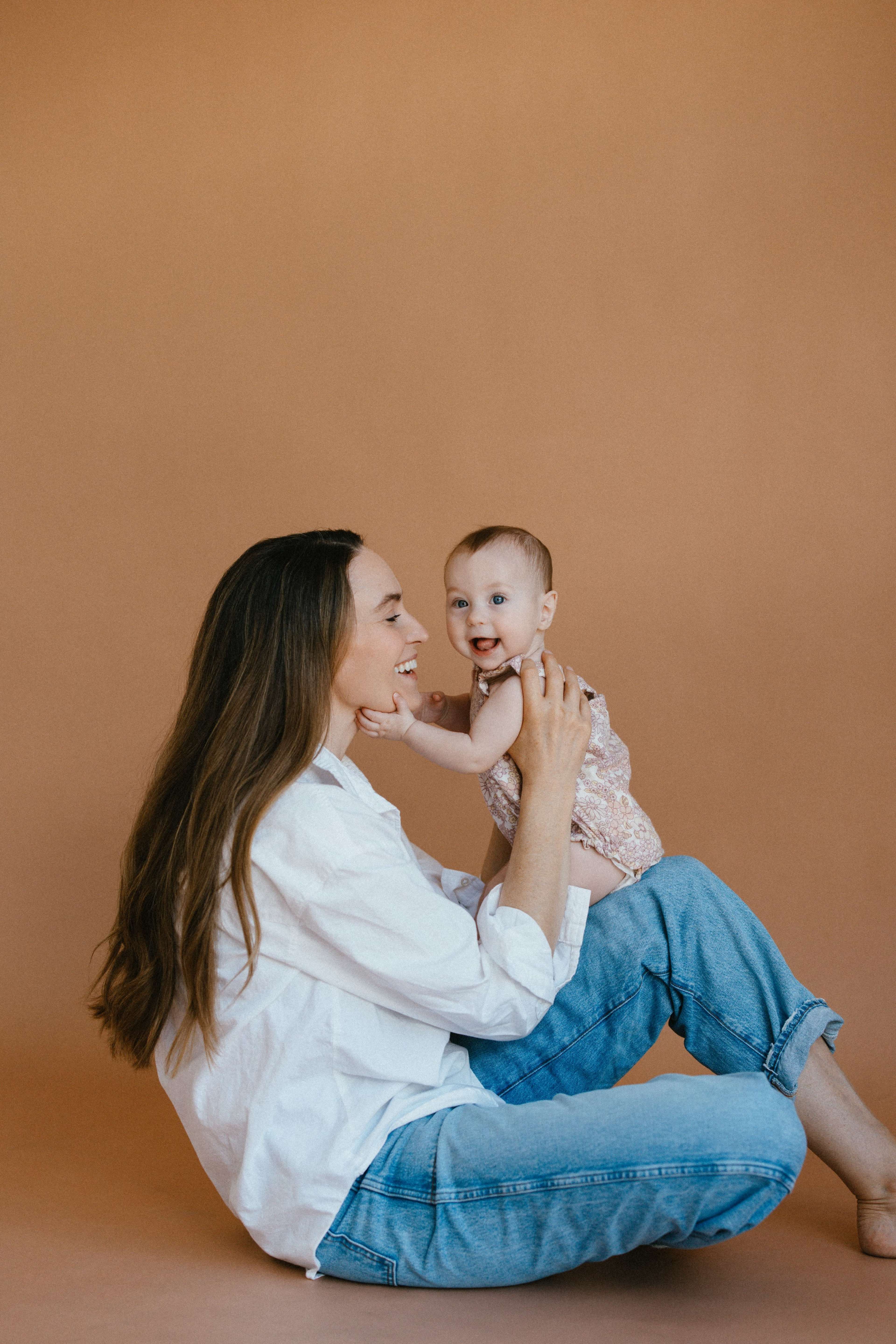 A woman sits on the floor with her legs bent, holding a smiling baby in a floral romper against a solid brown backdrop.